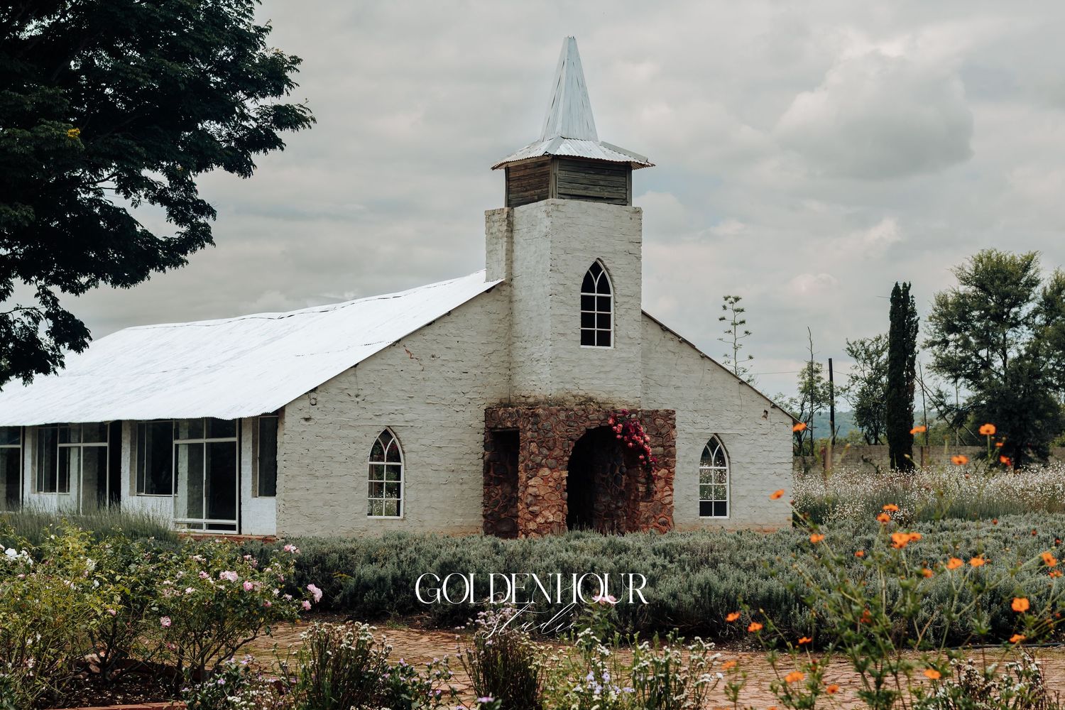 A rustic white church building stands in a wildflower garden under cloudy skies.