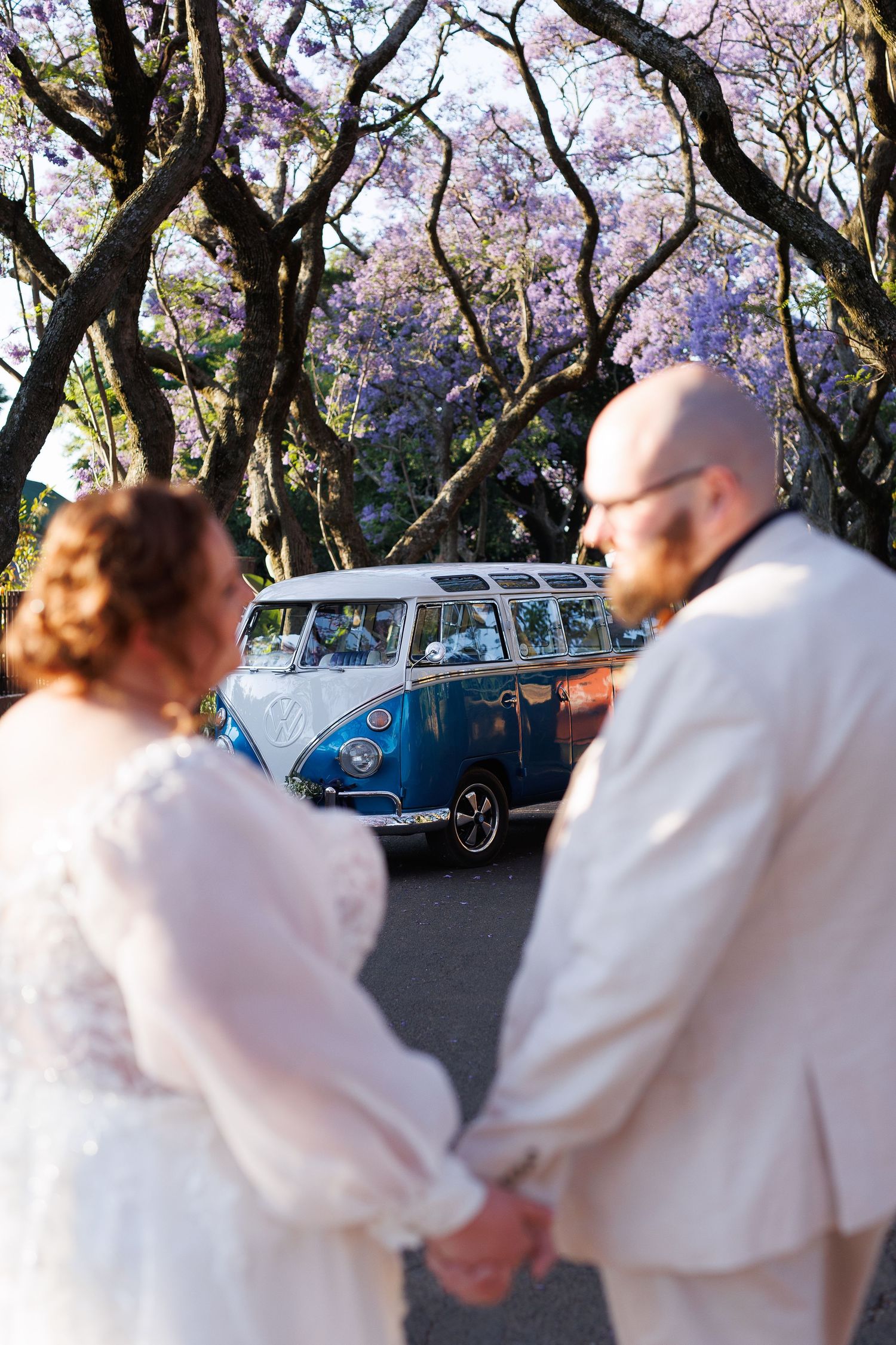 A couple holds hands near a classic blue and white Volkswagen van under blooming purple jacaranda trees.