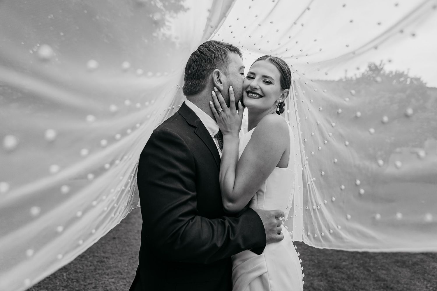 Black and white wedding photo capturing an intimate moment between newlyweds with a flowing veil in the wind.