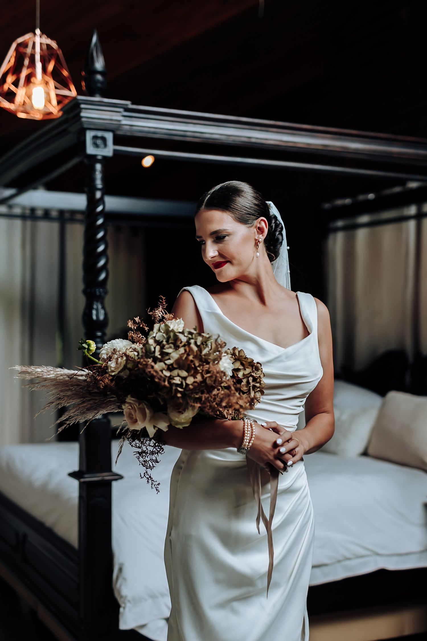 A bride in an elegant white dress holds a rustic dried flower bouquet in an industrial-style indoor setting.