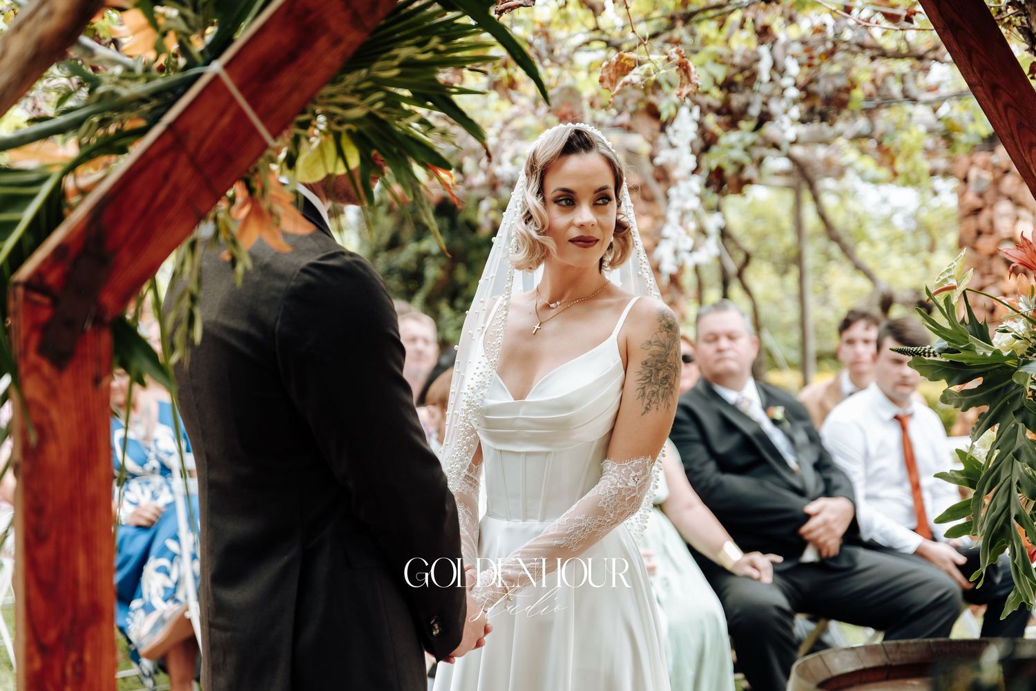 Wedding ceremony takes place under rustic wooden arbor decorated with greenery.