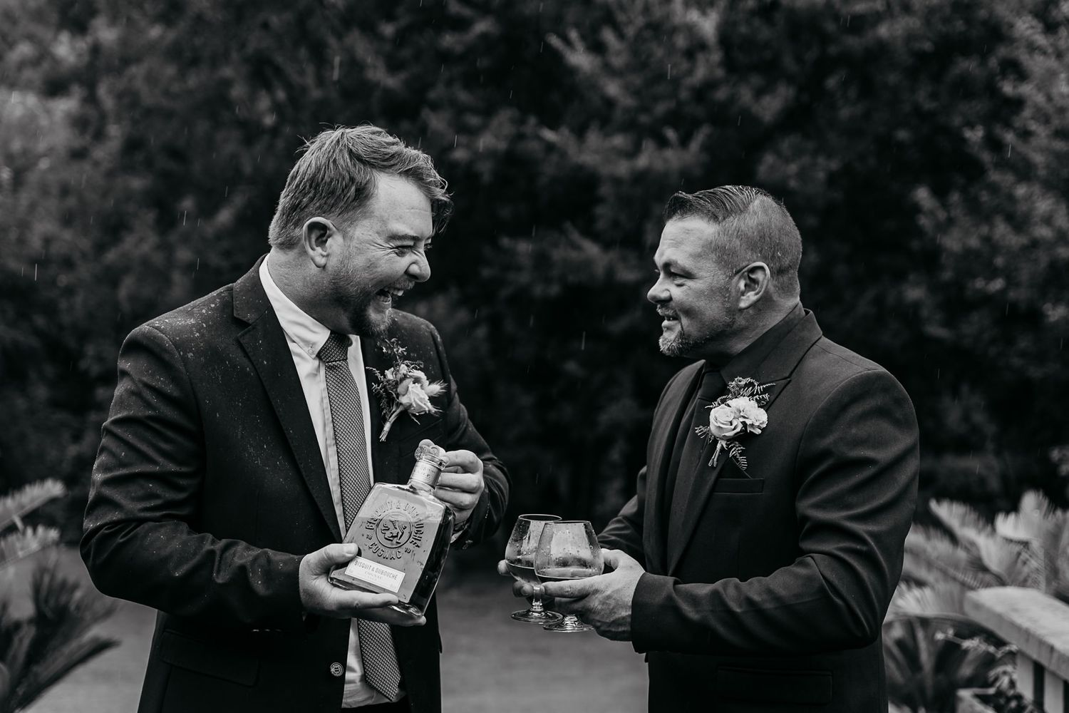 Two people in formal attire share a celebratory toast with champagne glasses outdoors in black and white photography.