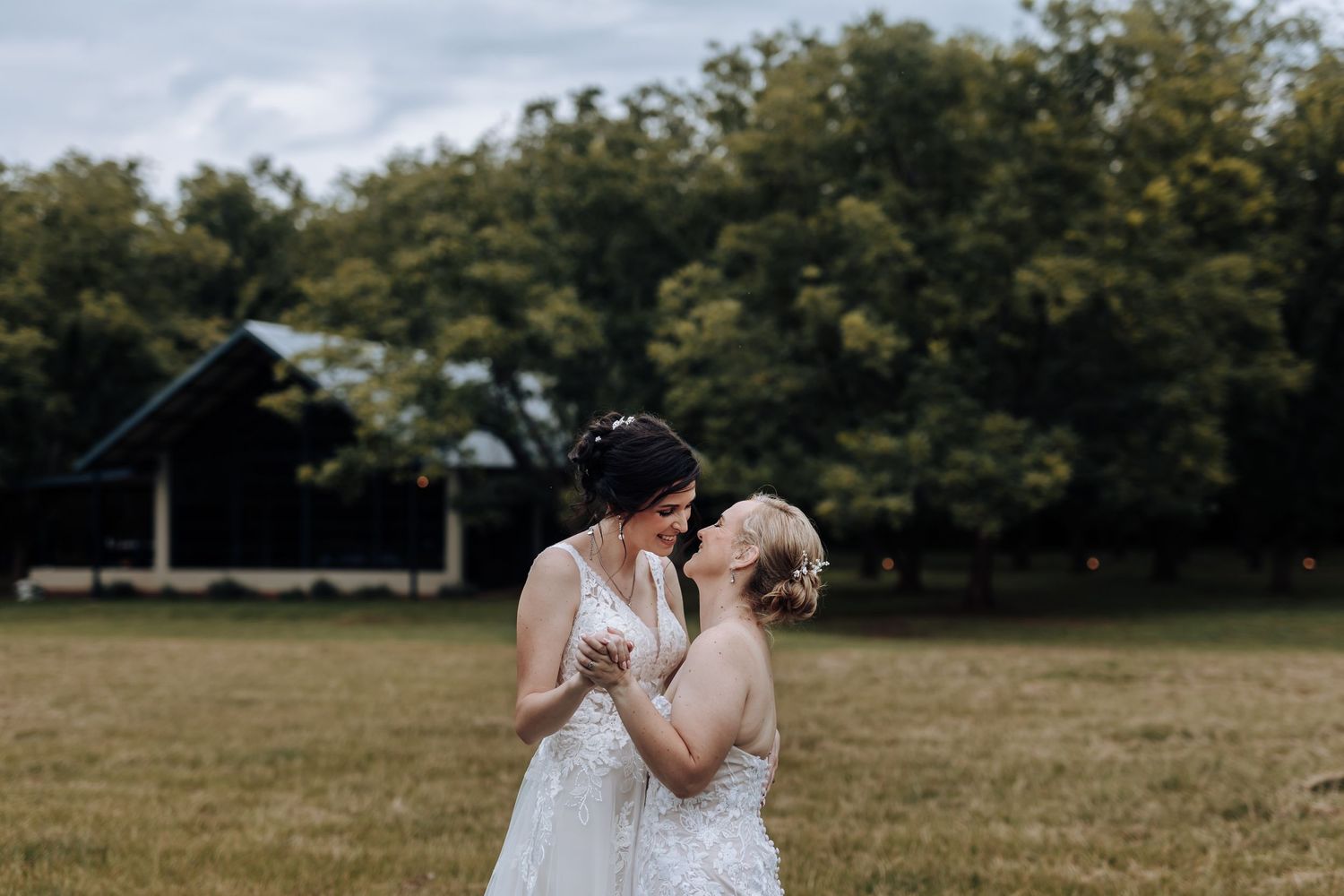 Two people in white dresses share a romantic moment in an outdoor field.