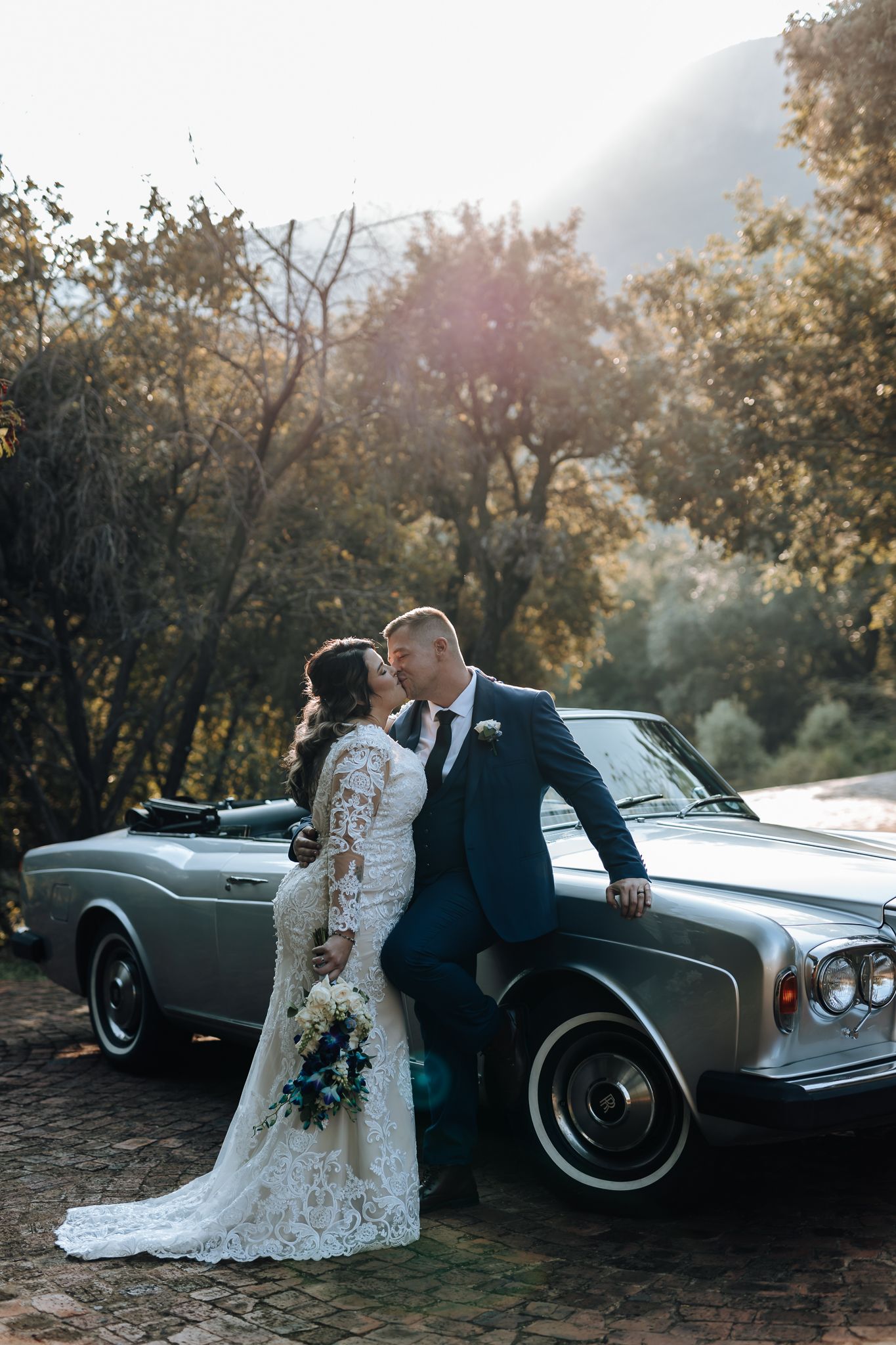 Romantic wedding portrait with couple kissing against classic car in mountain setting.