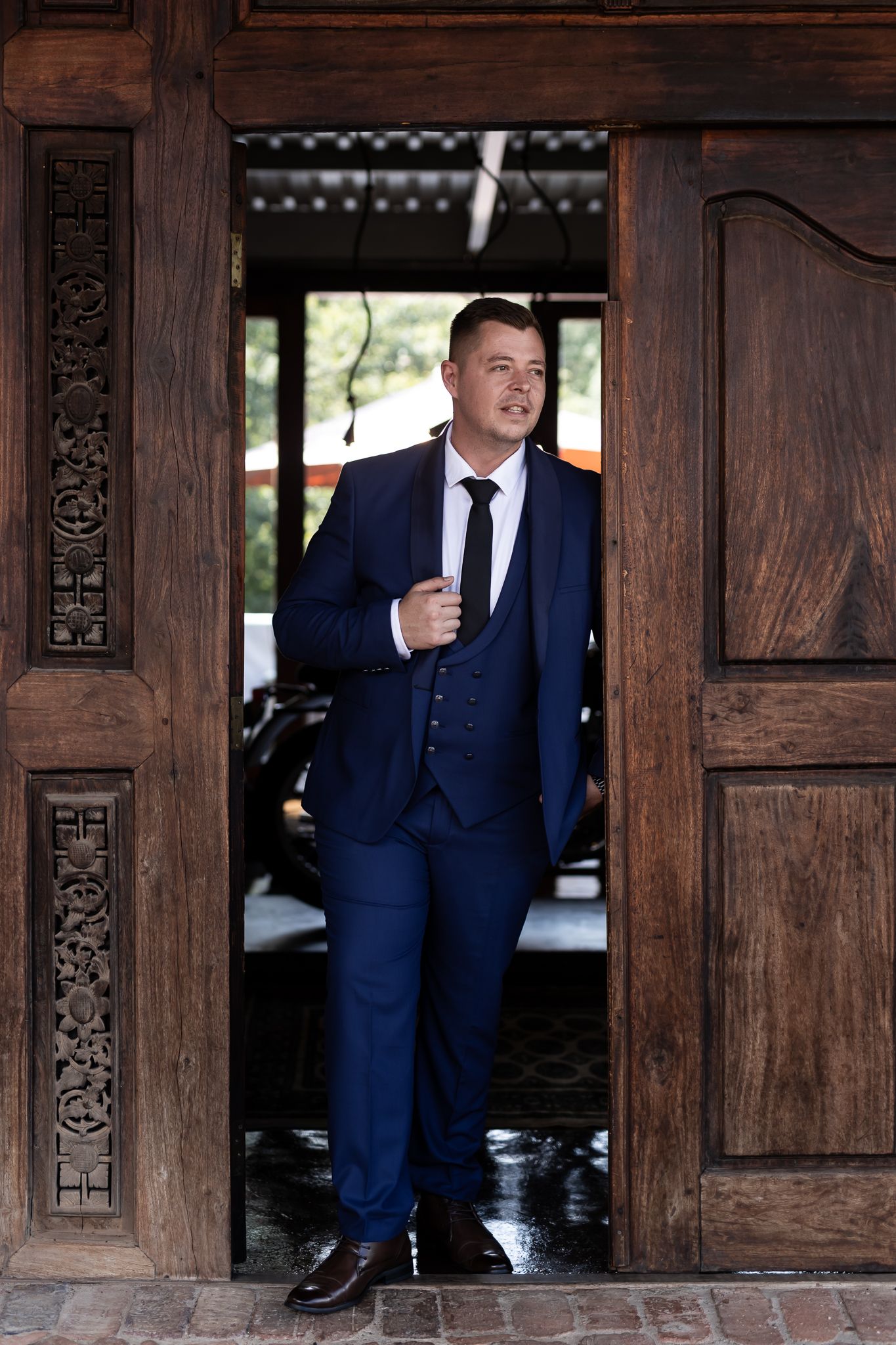 Man in navy blue three-piece suit posing confidently in wooden doorframe.