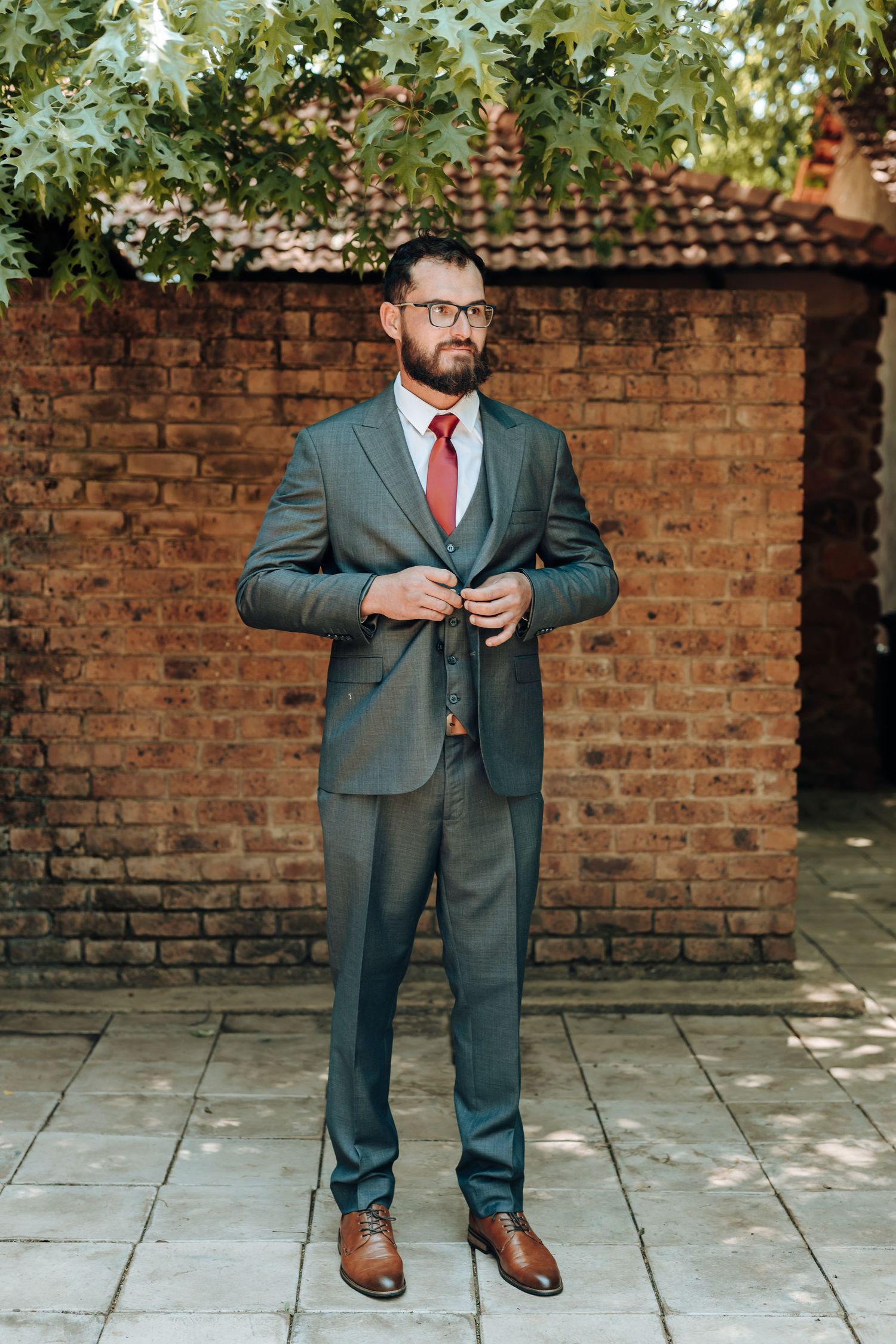 Full length portrait in grey suit and red tie against brick wall with foliage overhead.