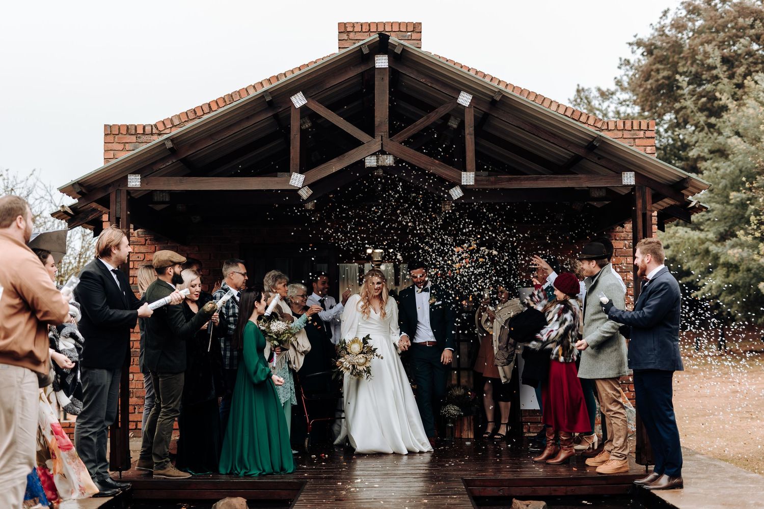 Wedding party and guests shower newlyweds with confetti outside rustic wooden pavilion.