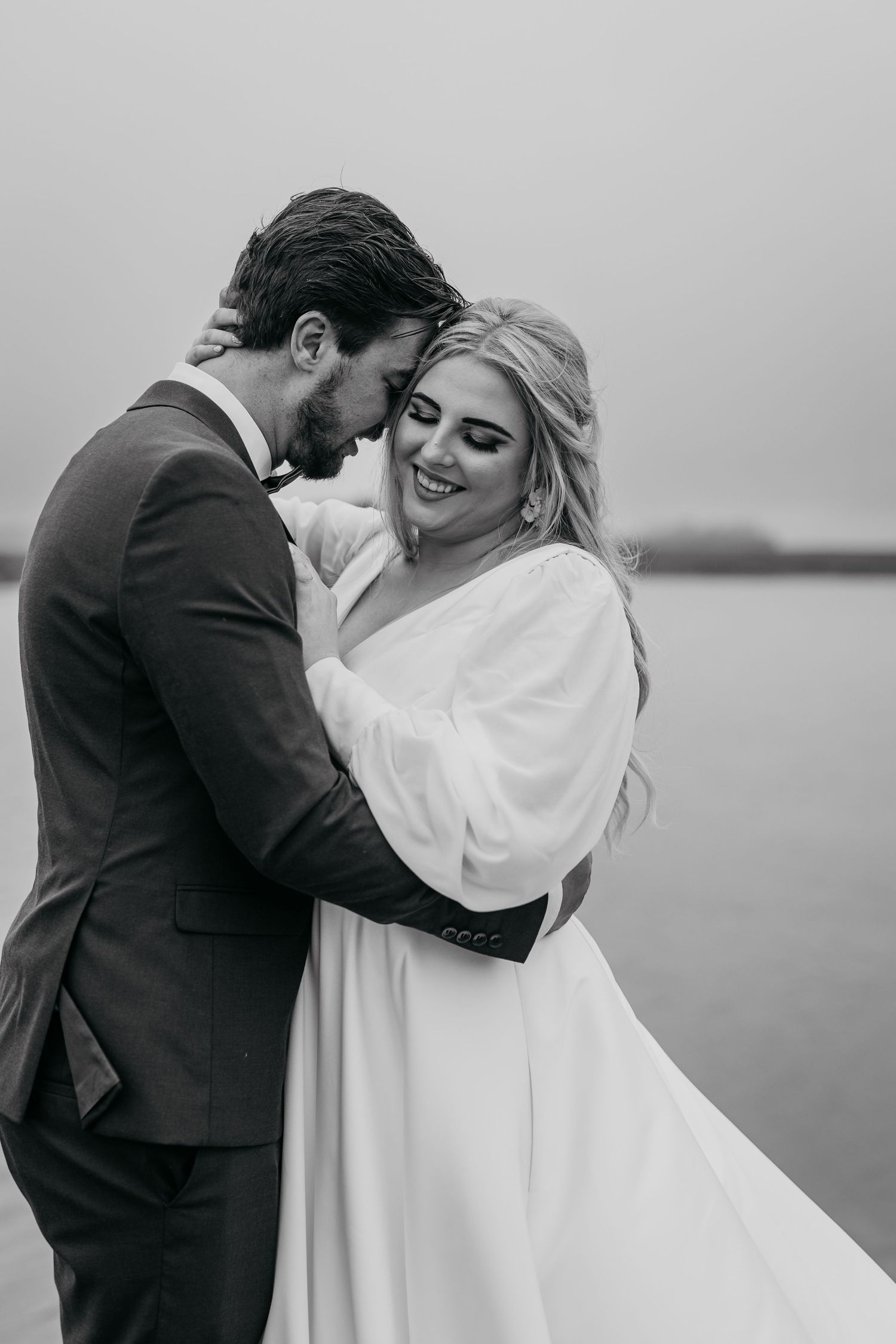 A romantic black and white wedding portrait captures an intimate moment between newlyweds by the water.