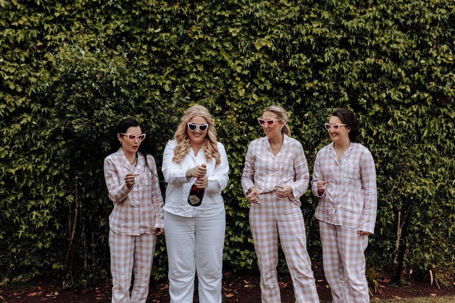 A group in pink and white checkered pajamas poses together against a hedge wall.