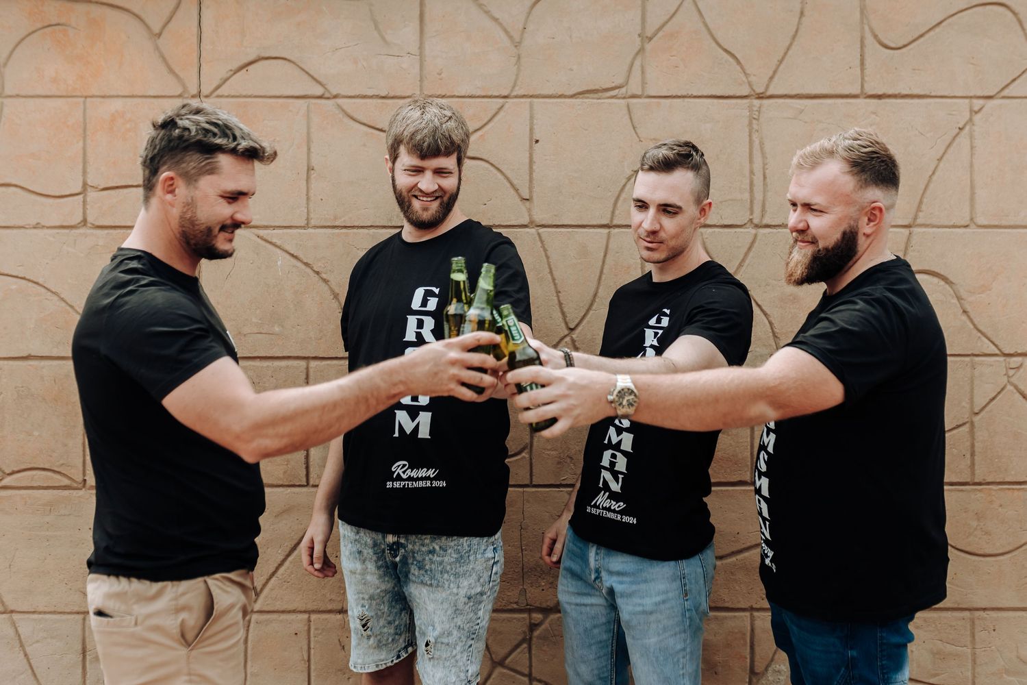 Friends in black t-shirts toasting with bottles against beige wall background.