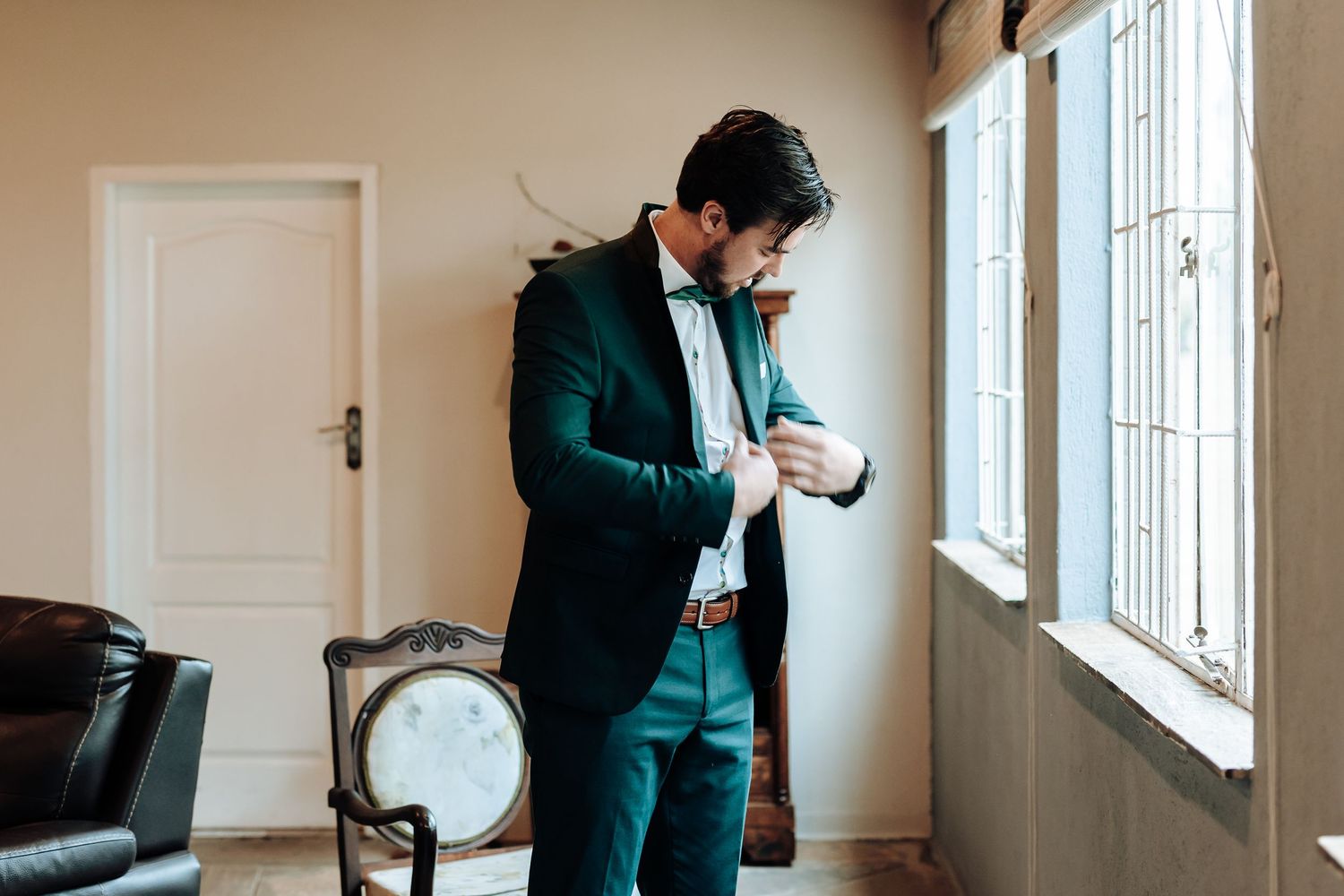 Emerald green suit being adjusted near a bright window with antique furniture in room.