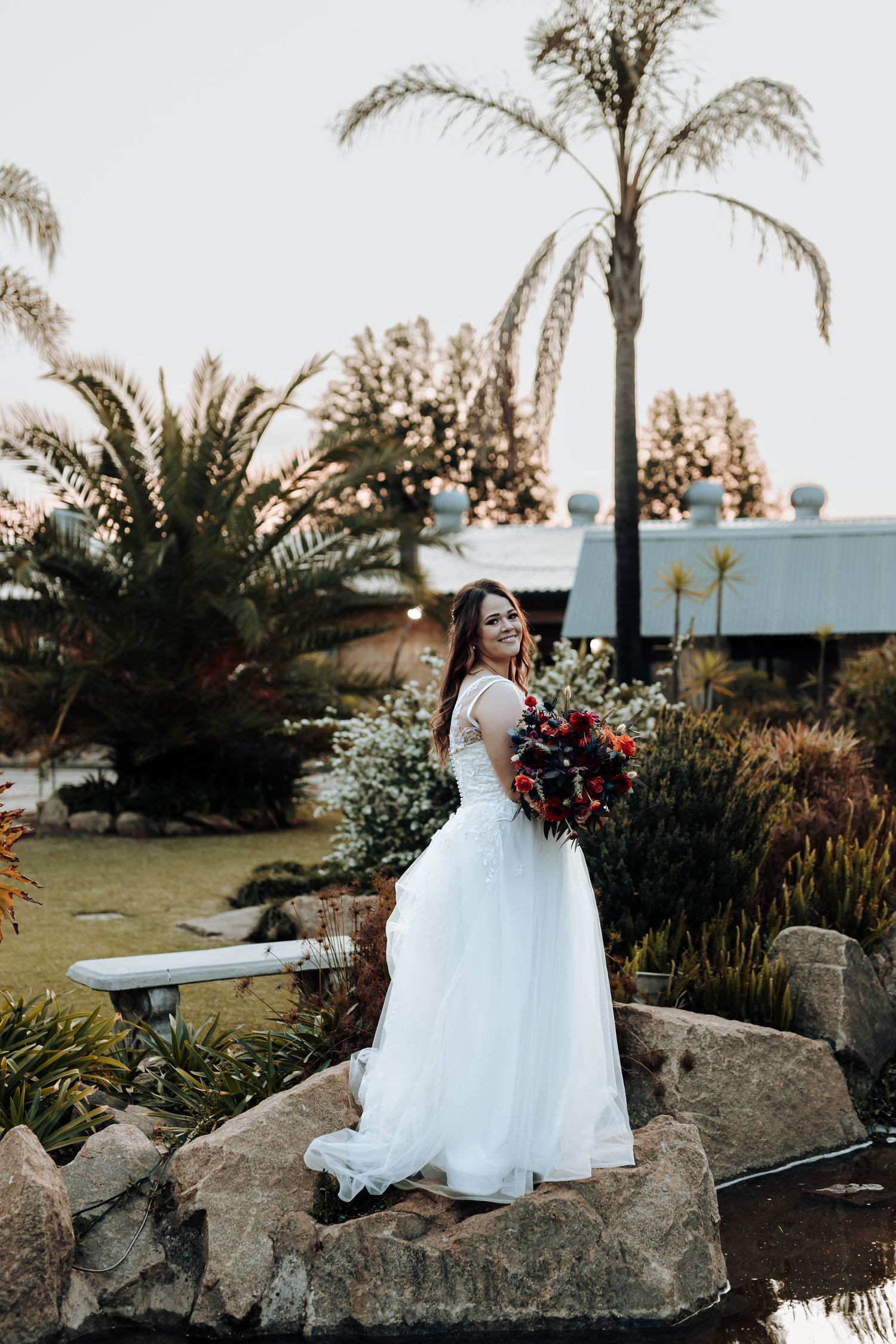 Bride in flowing white dress holds bouquet while posing on rock formation in garden setting.