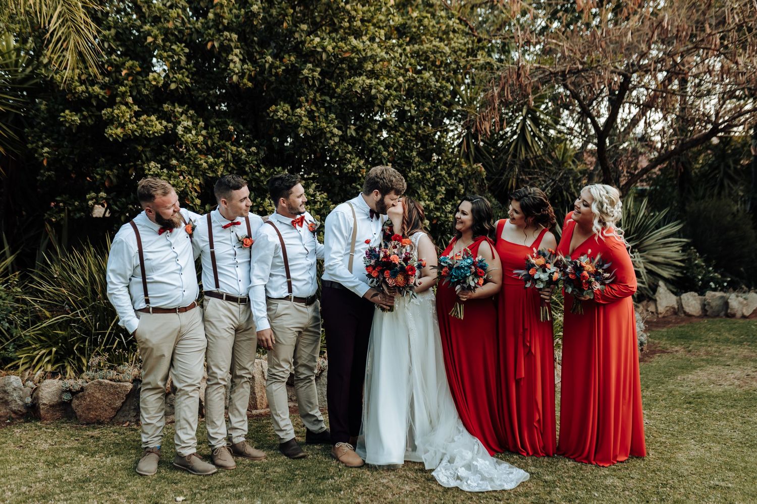 Wedding group photo with groomsmen in khaki pants and bridesmaids wearing vibrant red dresses.
