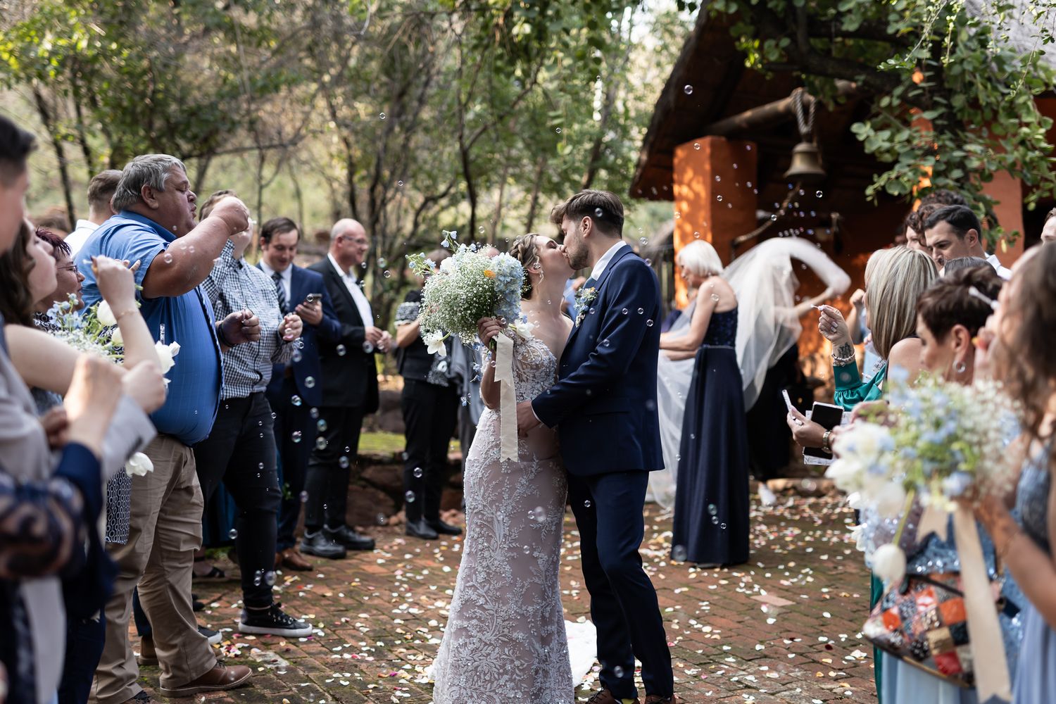Wedding guests throw confetti as newlyweds exit through outdoor garden pathway.