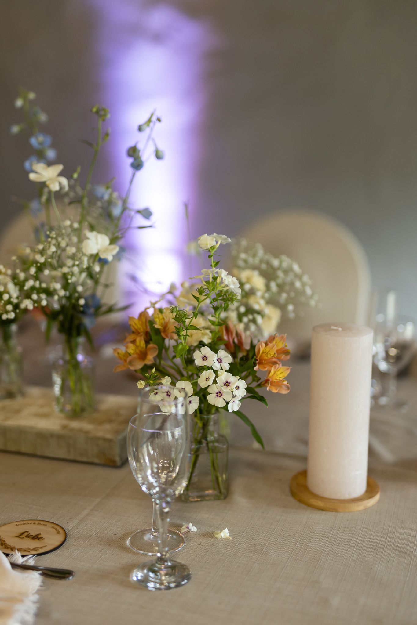 Delicate wildflower centerpiece with baby's breath in glass vase next to white candle on linen tablecloth.