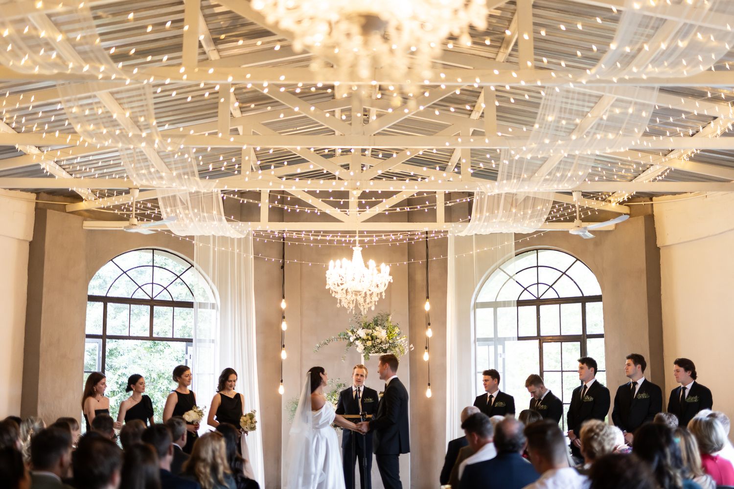 Wedding ceremony in a bright venue with crystal chandeliers and guests watching a couple exchange vows.