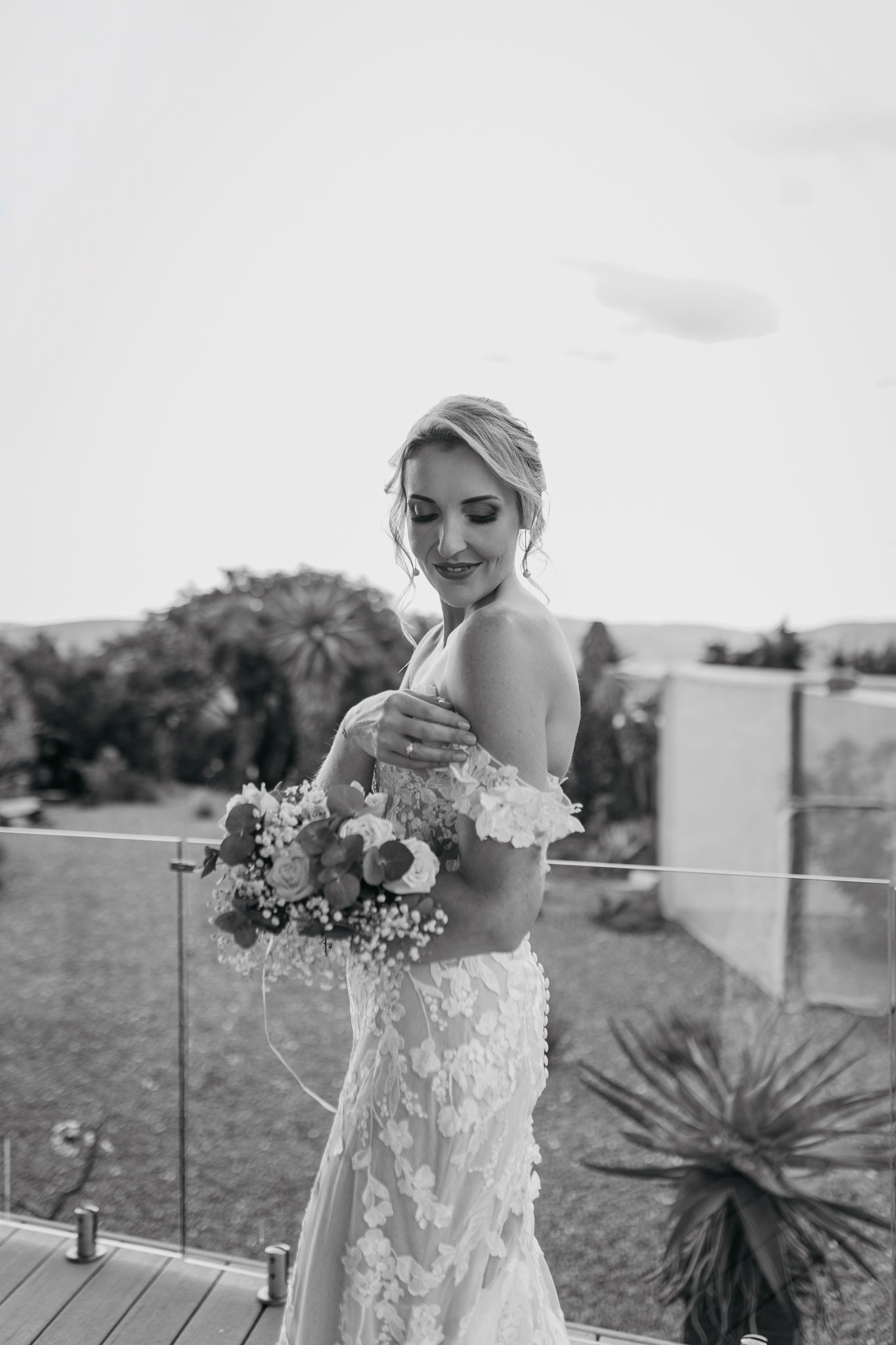 A bride in a lace wedding dress holds a bouquet while posing on a deck with mountain views.