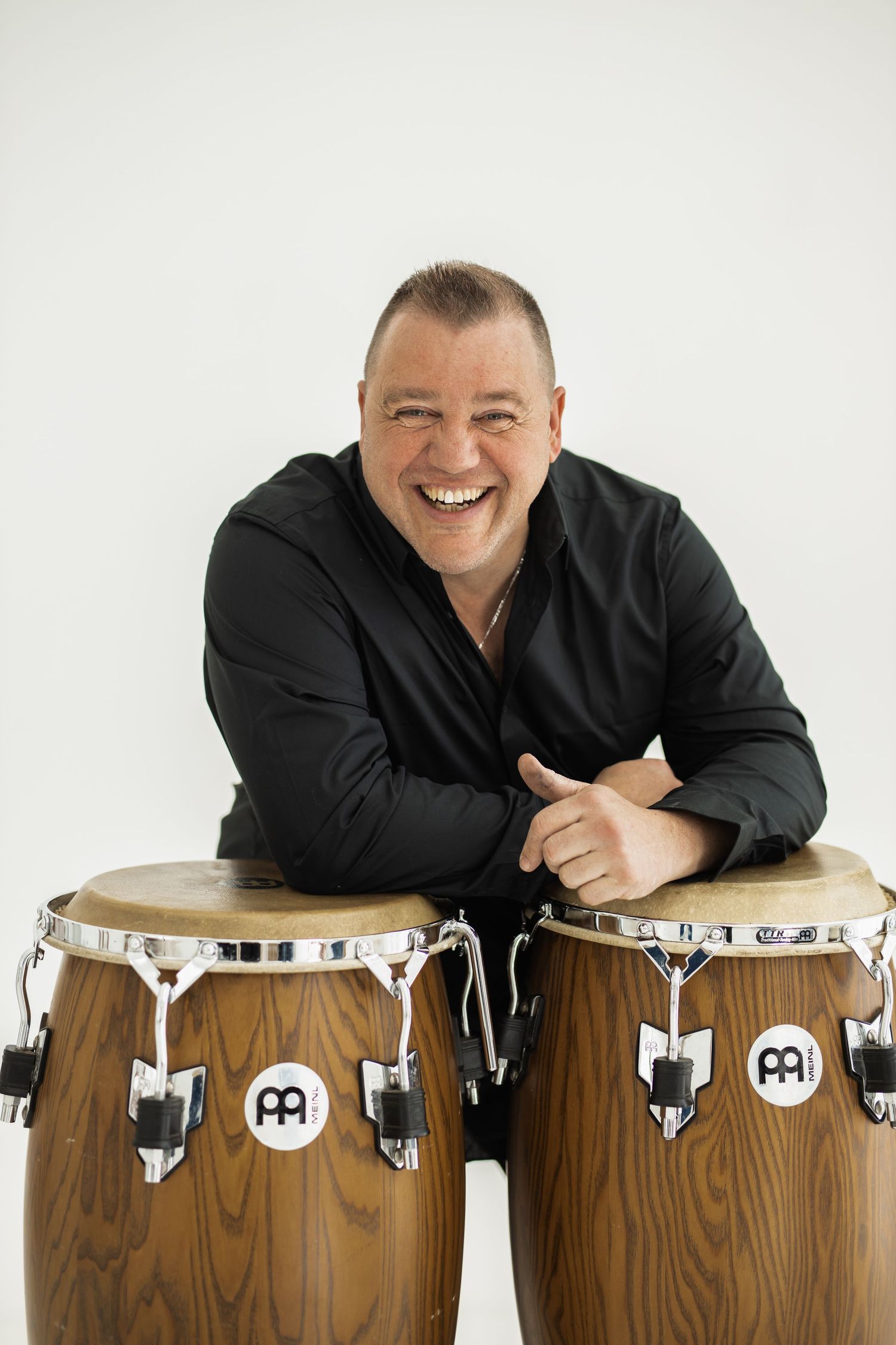 Professional musician wearing black shirt leans playfully on three wooden conga drums against white background.