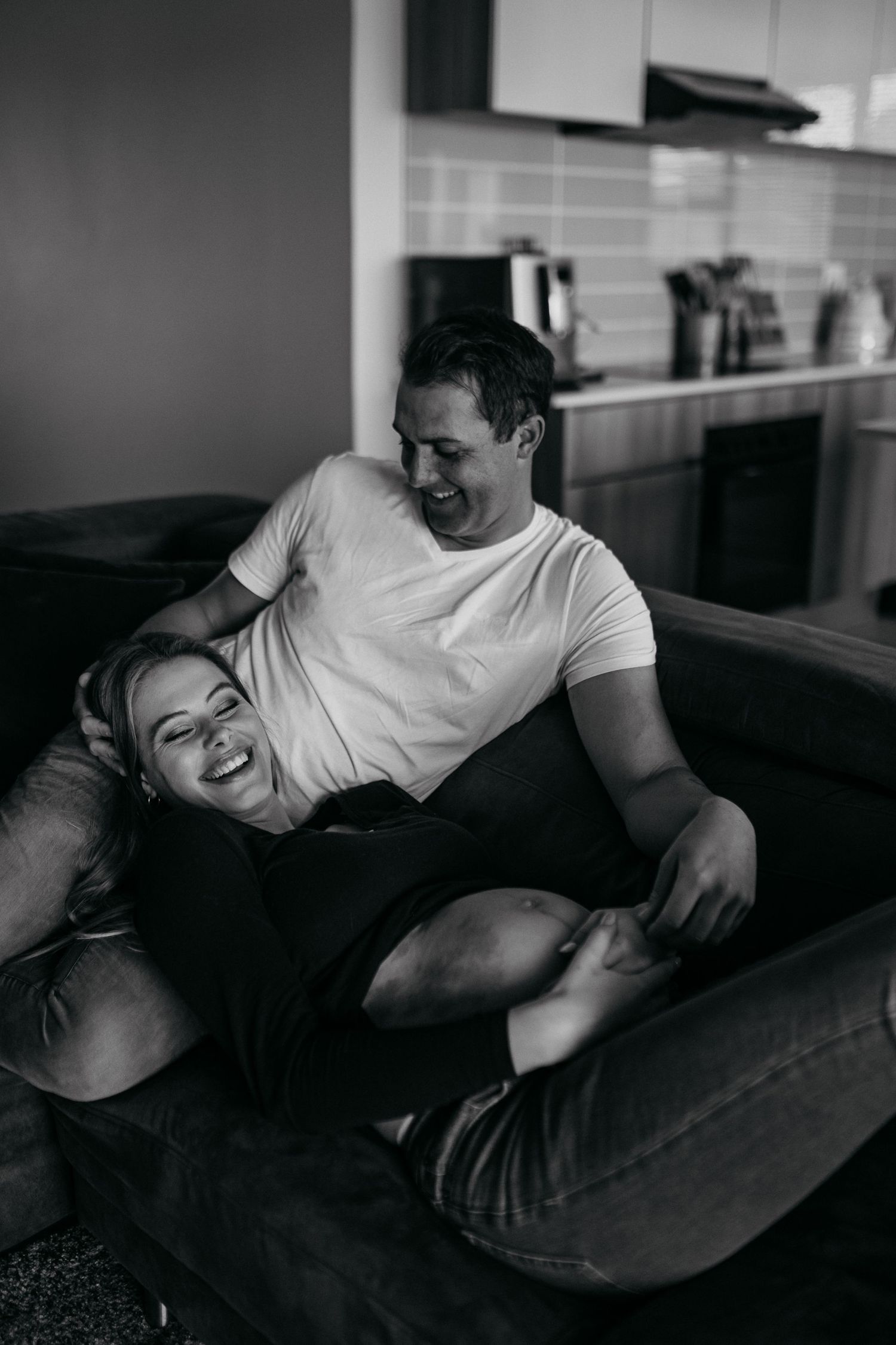 A candid black and white photo of a couple laughing together while relaxing on a dark couch in a kitchen setting.