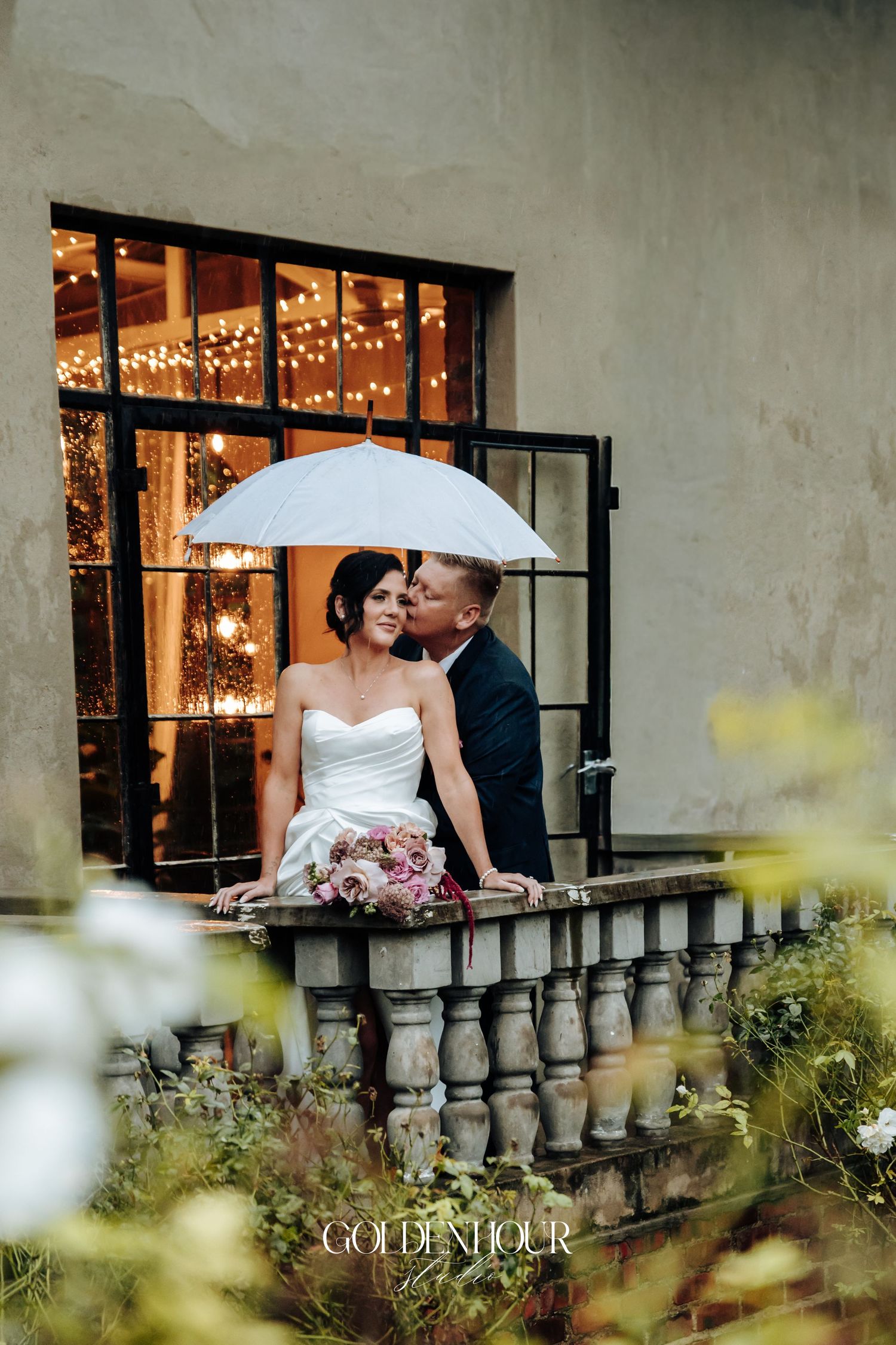 Bride and groom sharing a kiss under white umbrella on stone balcony with fairy lights.
