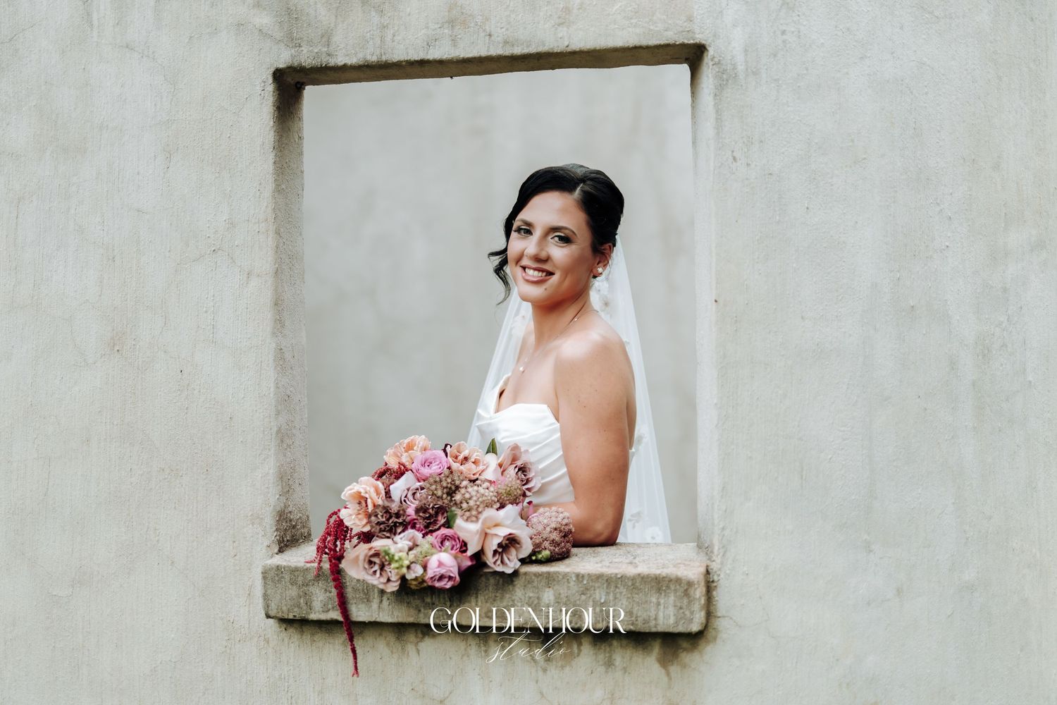 Smiling bride in white gown and veil holding a pink bouquet framed in a stone window alcove.