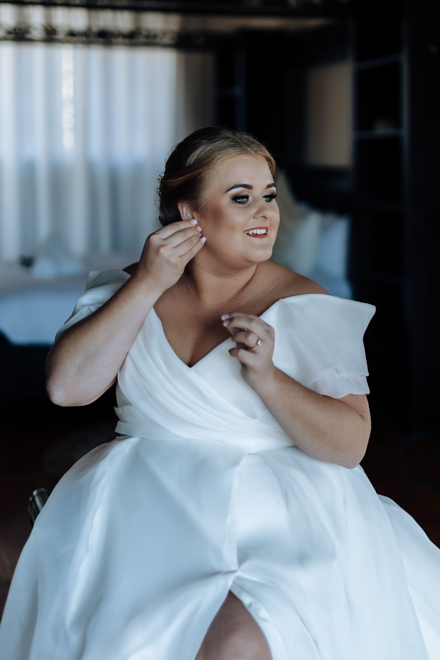 Smiling bride in white off-shoulder gown adjusting earring while seated, wedding day preparation photo.