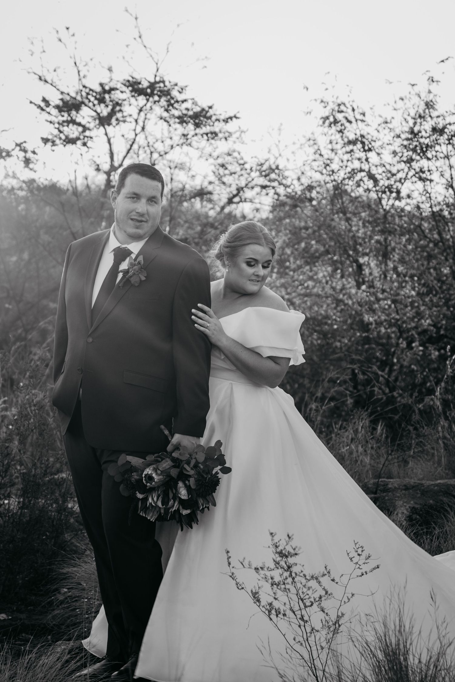 Black and white photo of a bride in an off-shoulder gown holding a bouquet, standing with a groom in a suit outdoors.
