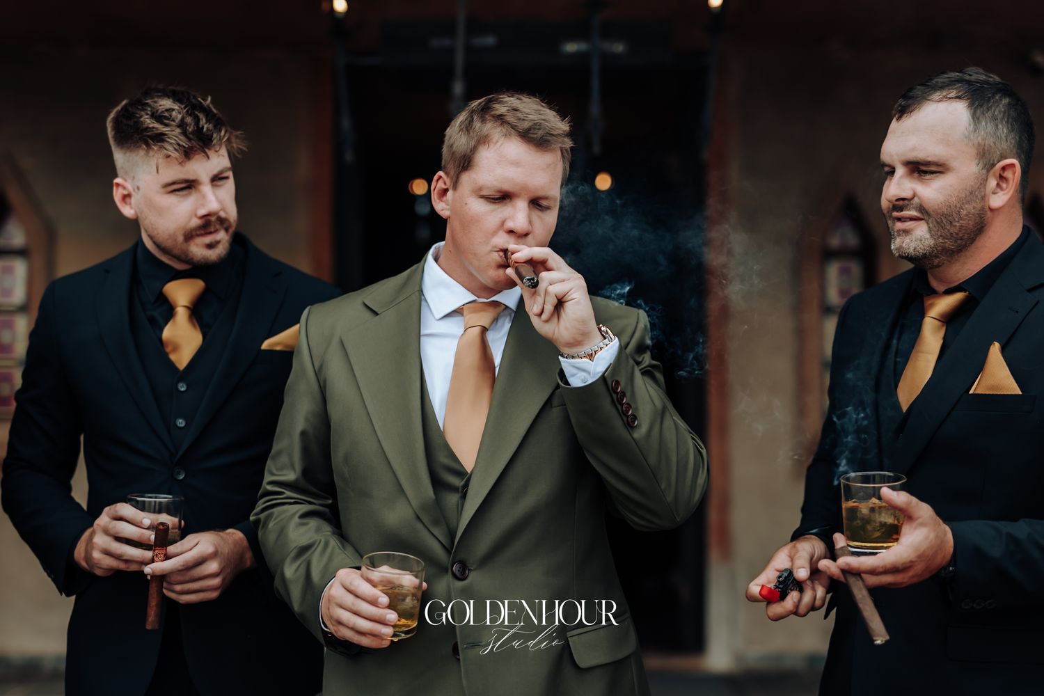 Three groomsmen in suits enjoy cigars and drinks outdoors, groom in olive green suit centered.