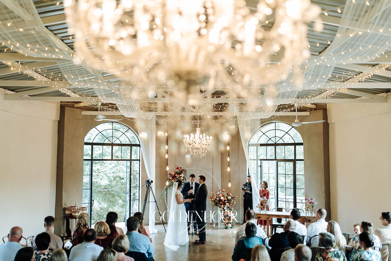 Elegant wedding ceremony under glittering chandeliers in a bright white venue with arched windows.