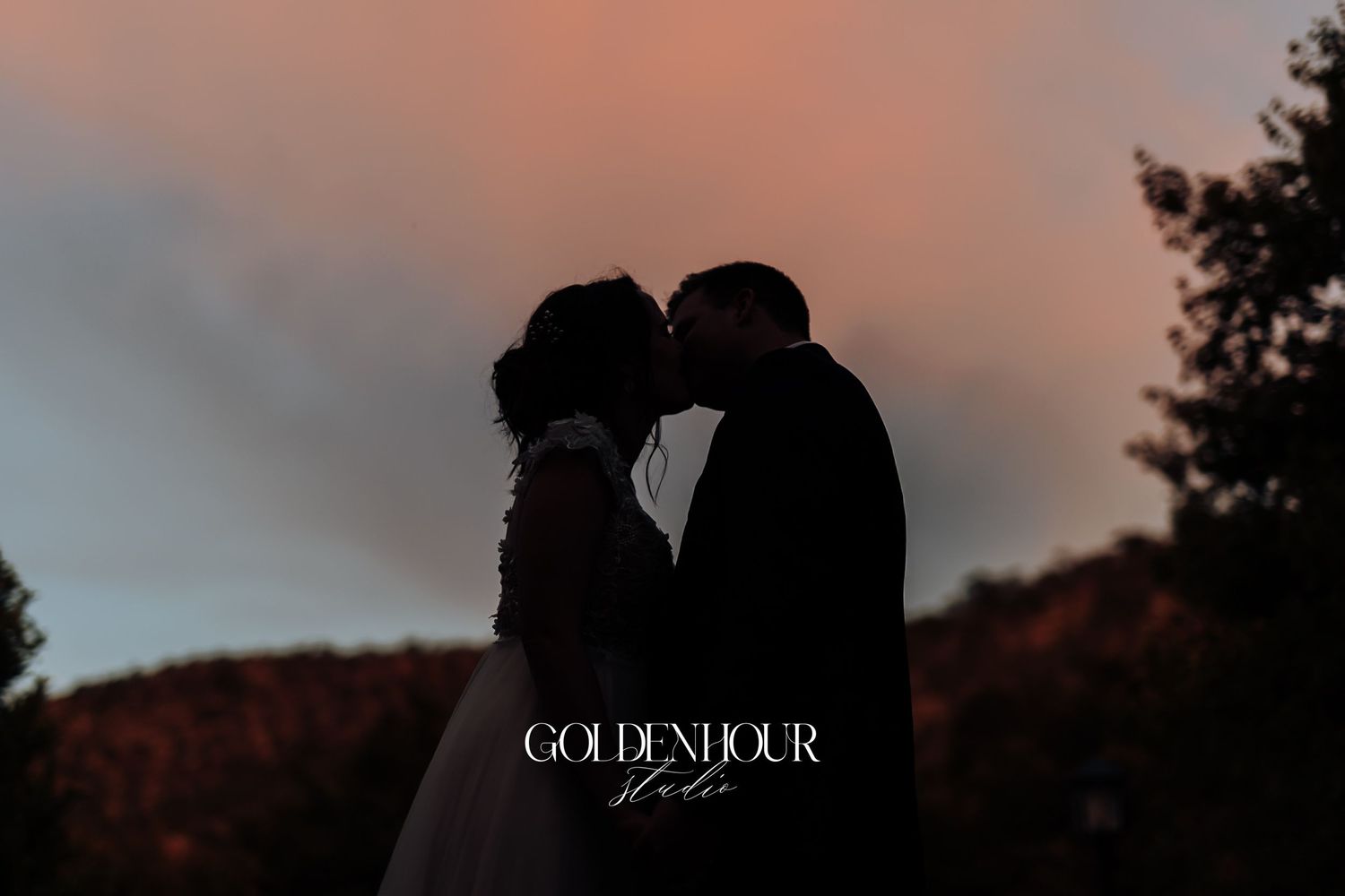 Silhouette of bride and groom sharing a kiss against a dramatic sunset sky.