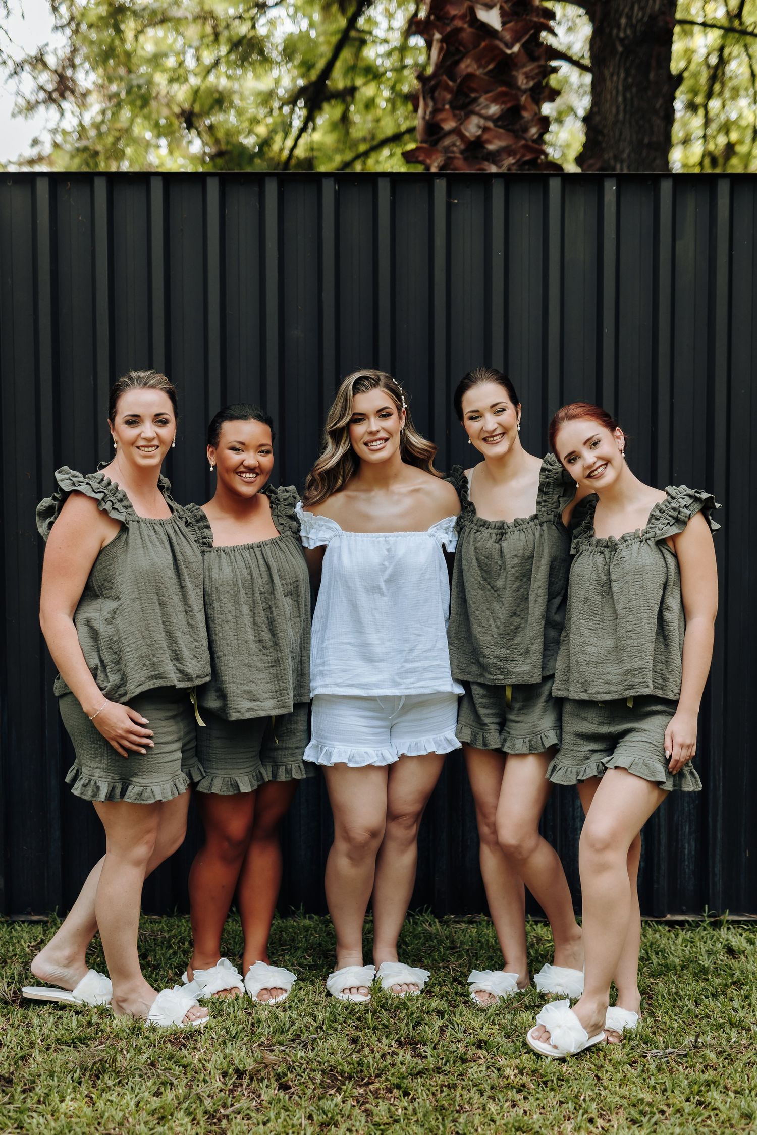 Bridesmaids in olive green tops and white shorts pose together on grass in front of black fence.