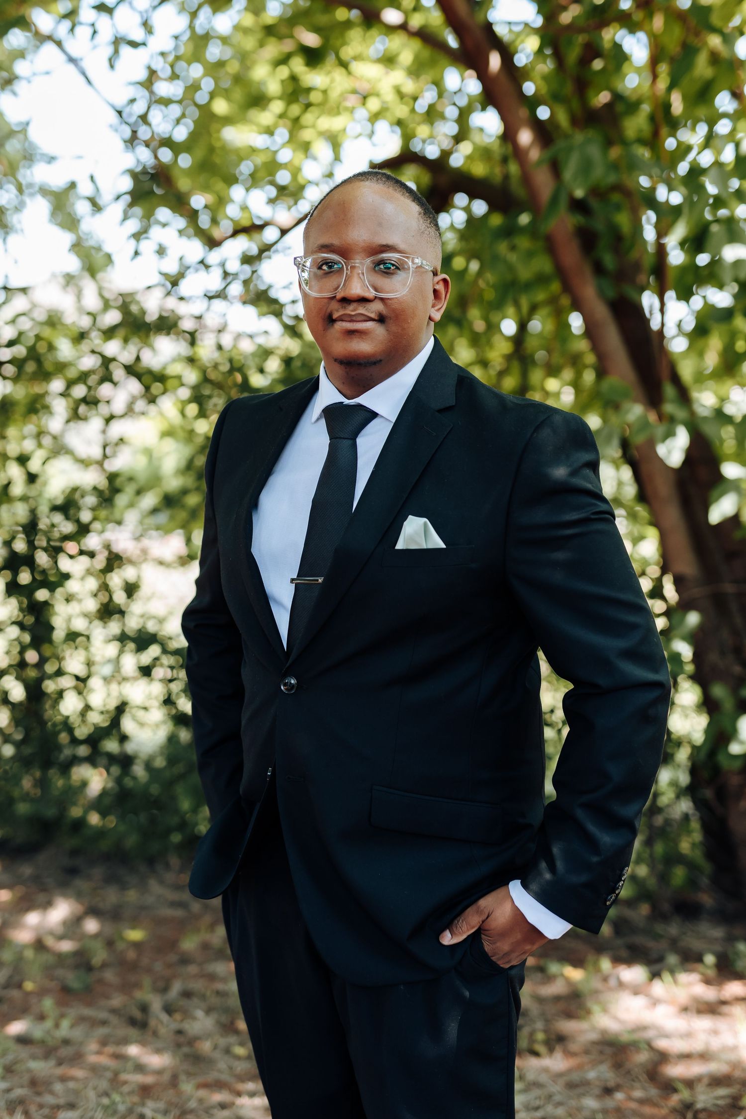 Person in formal black tuxedo and white shirt standing confidently in outdoor garden setting.