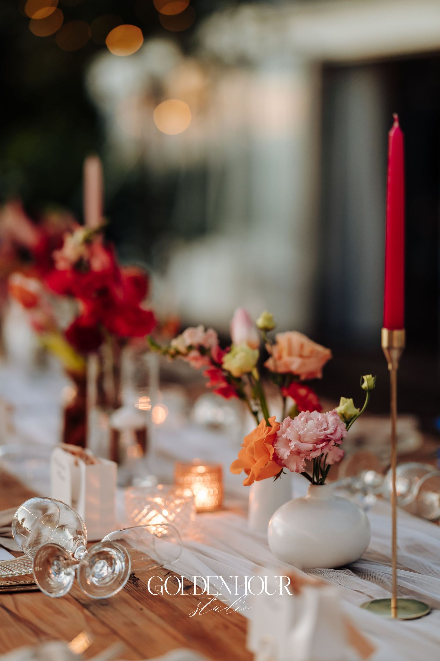 Elegant wedding reception table setting with red and pink floral centerpieces, white vases, and glowing candles.