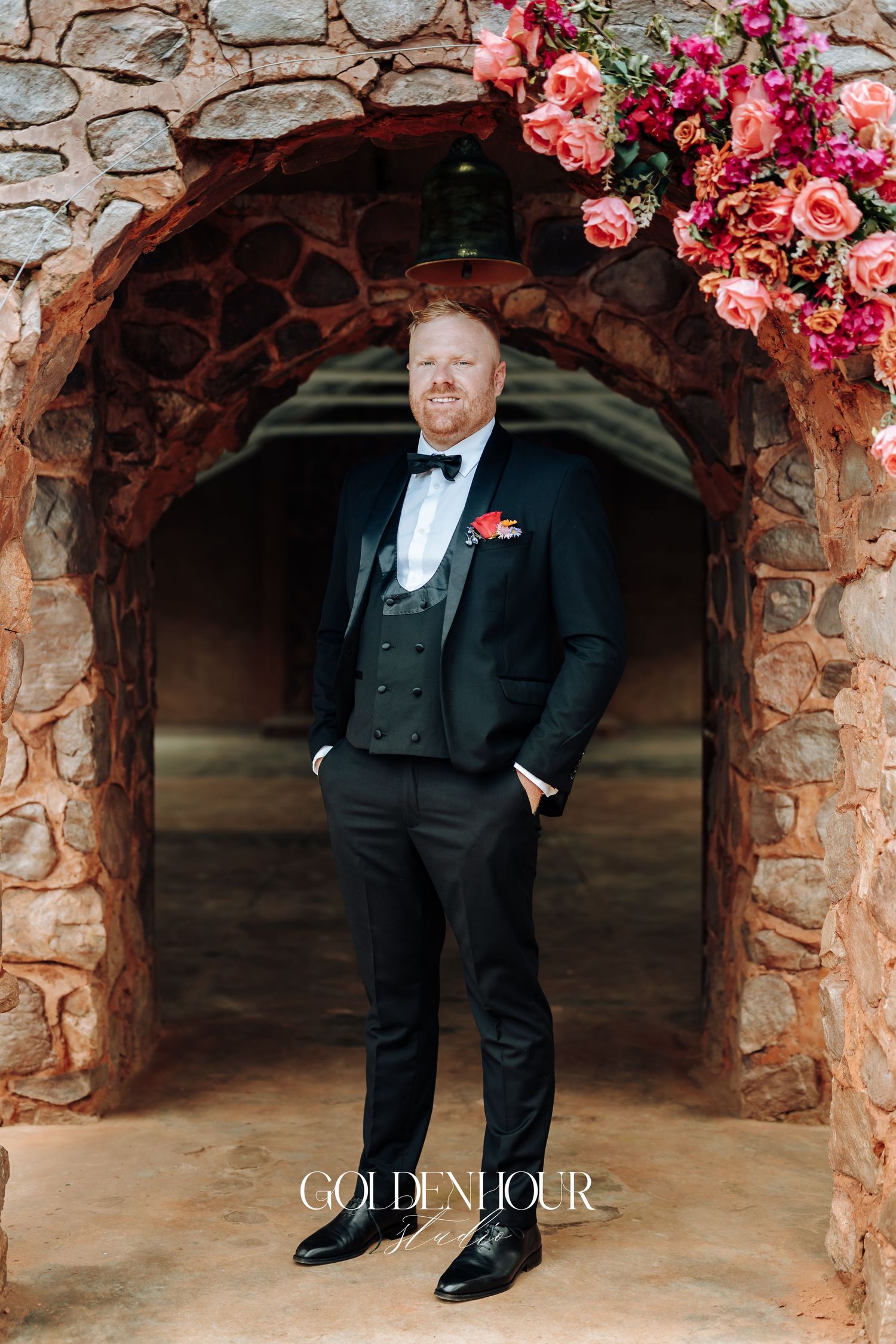 Person in formal black tuxedo standing in stone archway decorated with vibrant pink and coral floral arrangements.