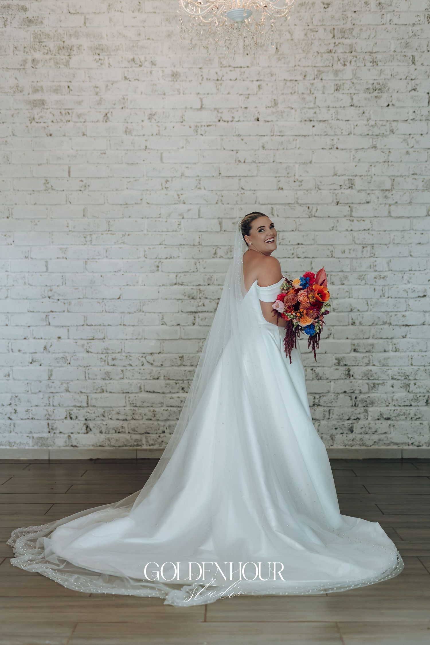 A bride in a flowing white gown holds a vibrant autumn bouquet while posing against a white brick wall.