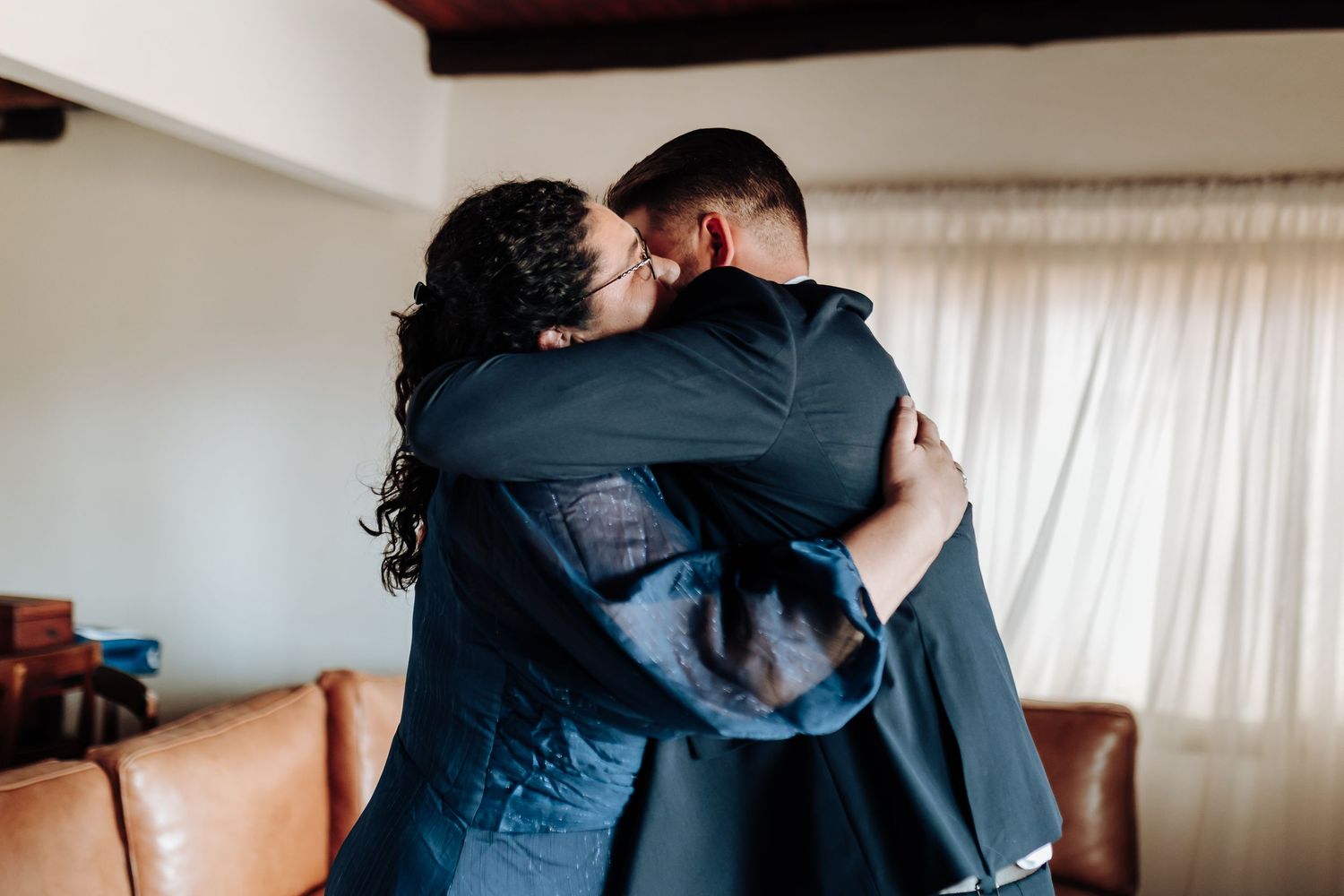 Two people embrace in an emotional hug wearing dark clothing in a room with a brown leather couch and white curtains.