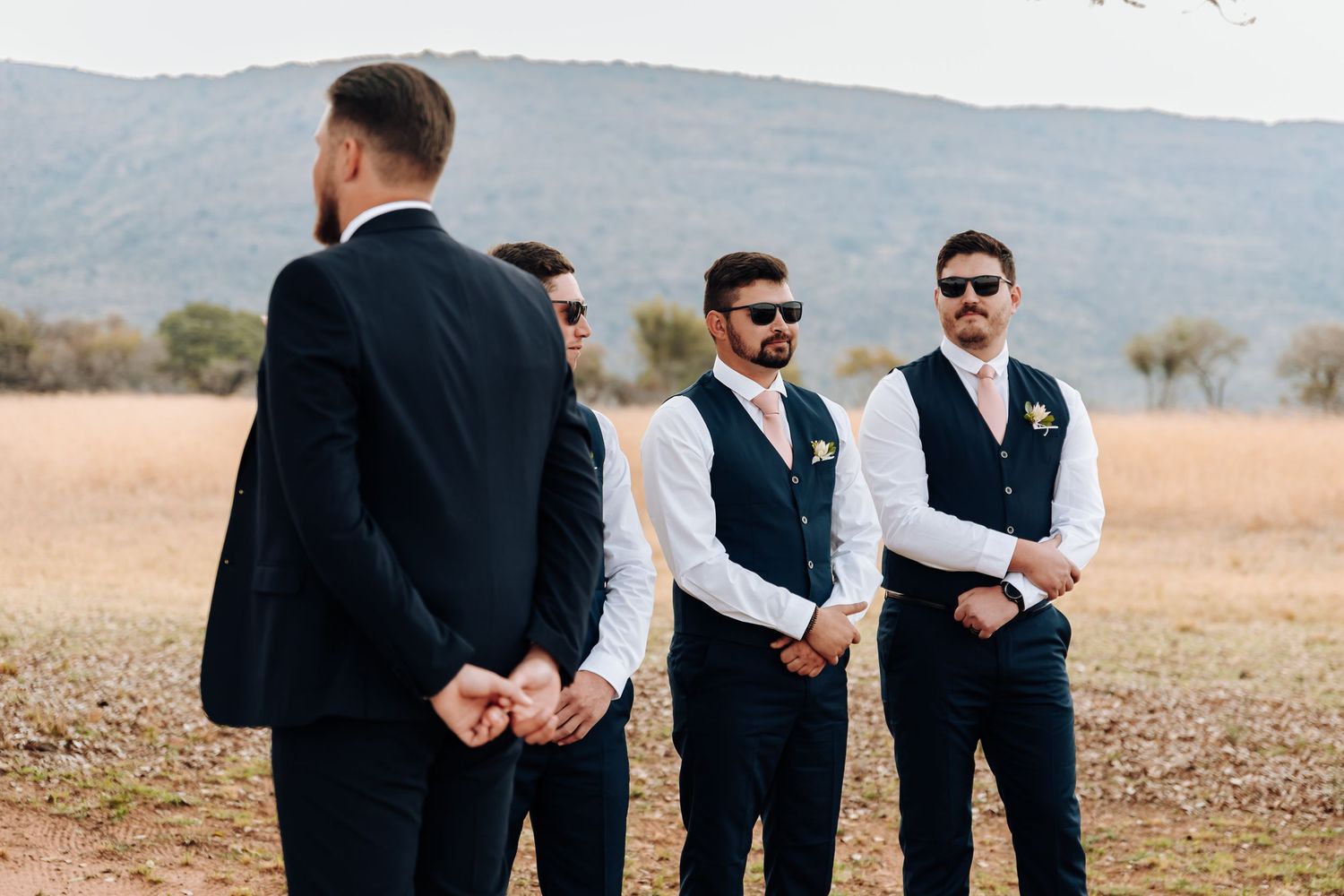 Groomsmen in navy vests and dress shirts stand in an open field with mountains visible in the background.