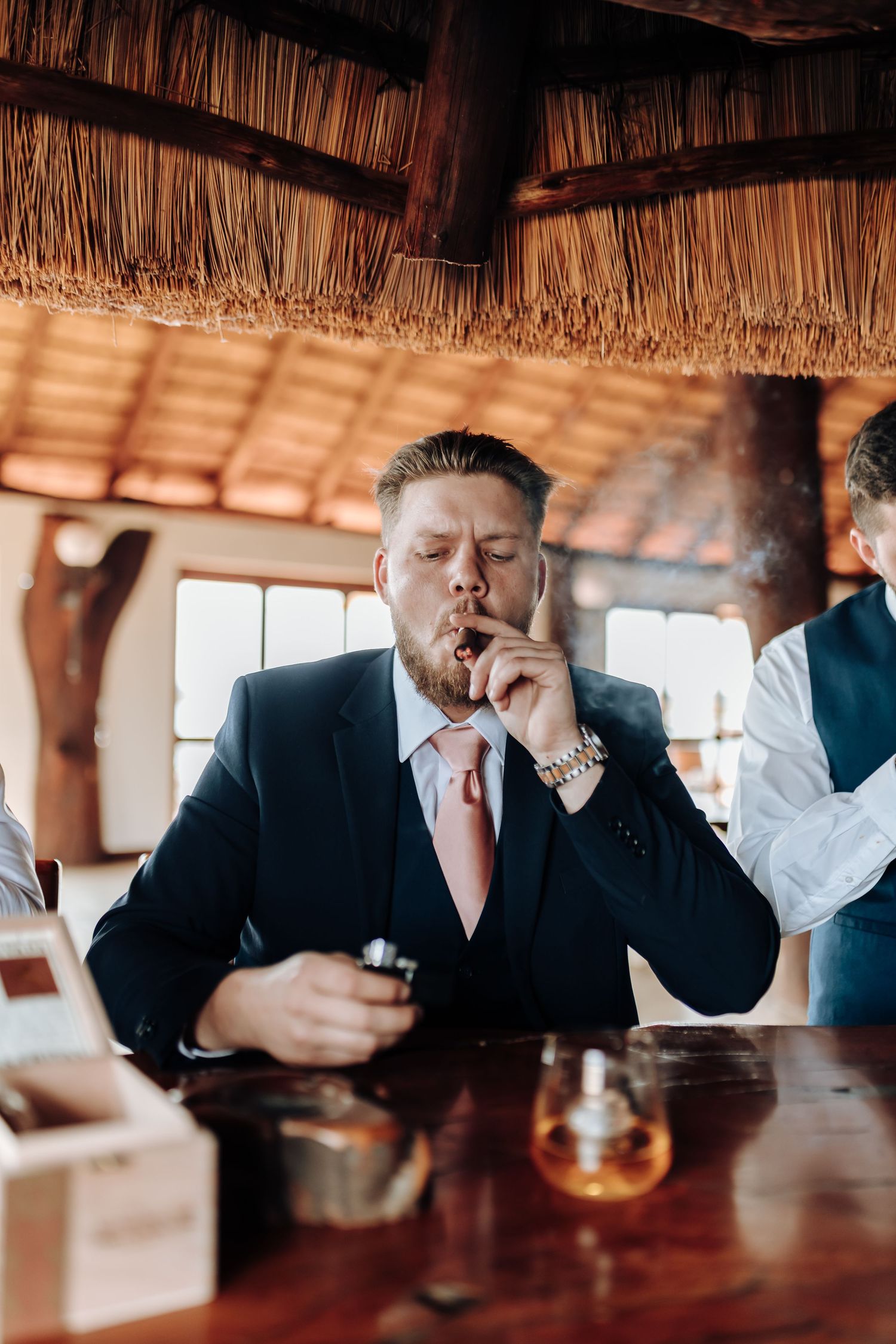 A person in a suit smoking a cigar at a bar under a thatched roof with drinks on the table.
