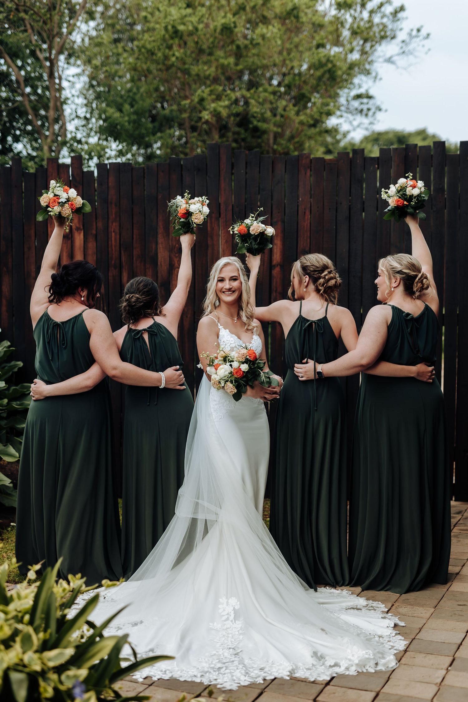 Bridal party in black dresses and white gown pose with their backs turned on stone patio showing coordinated arm gestures.