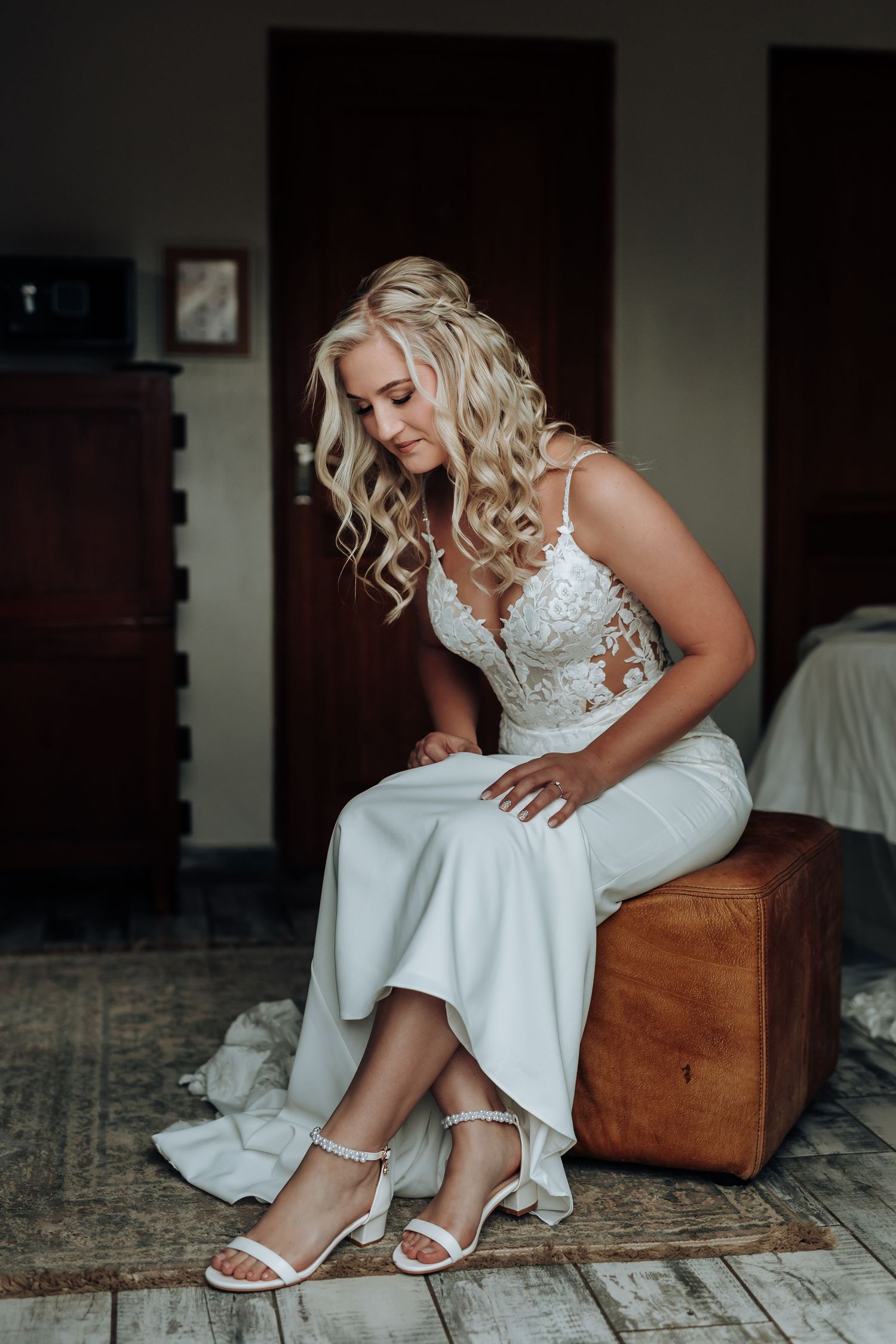 Close-up shots of a bride in a white wedding dress putting on white sandals while sitting on a brown leather chair.