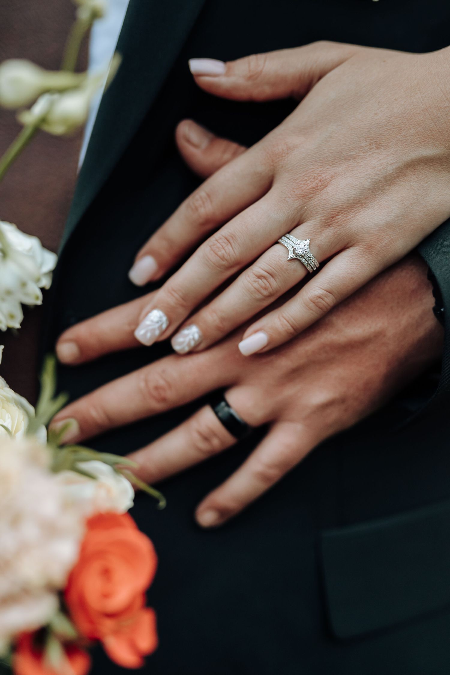 Close-up shot of hands wearing wedding rings with orange flowers and white floral decor against a dark background.