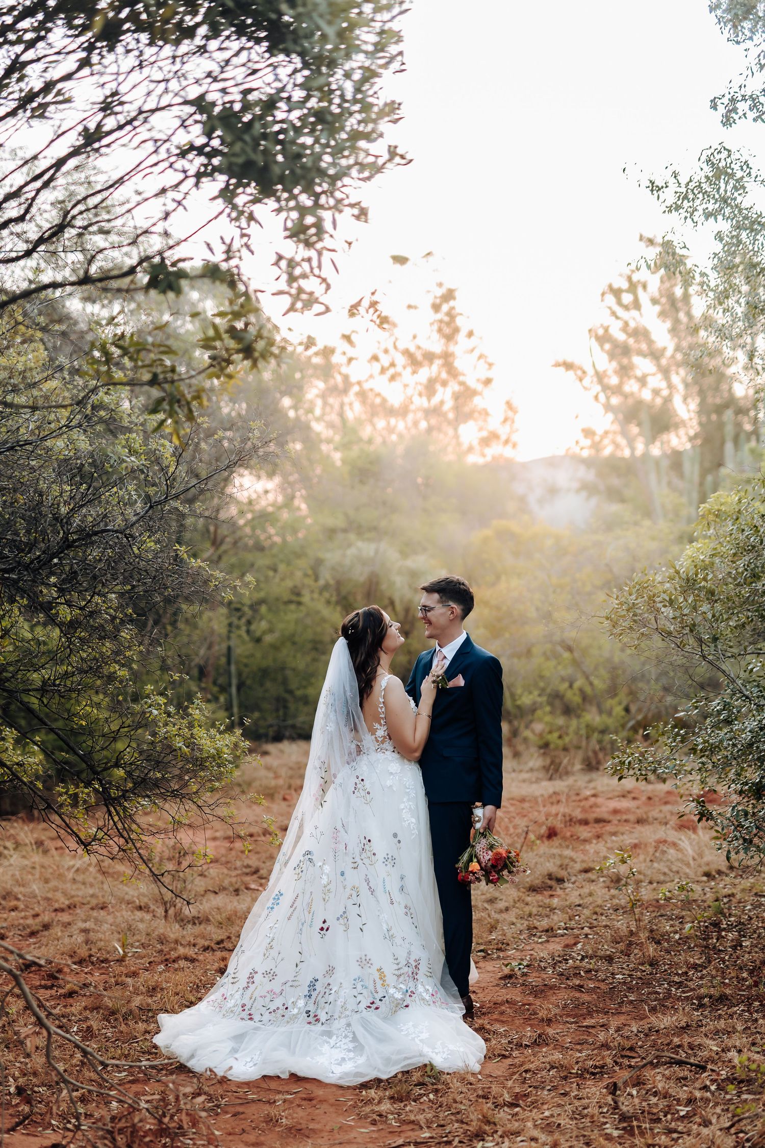 A romantic wedding couple shares an intimate moment in a sunlit forest setting with a flowing lace gown.