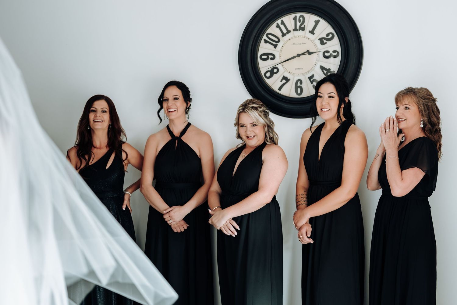 A group in black dresses poses against a white backdrop with a vintage clock on the wall.