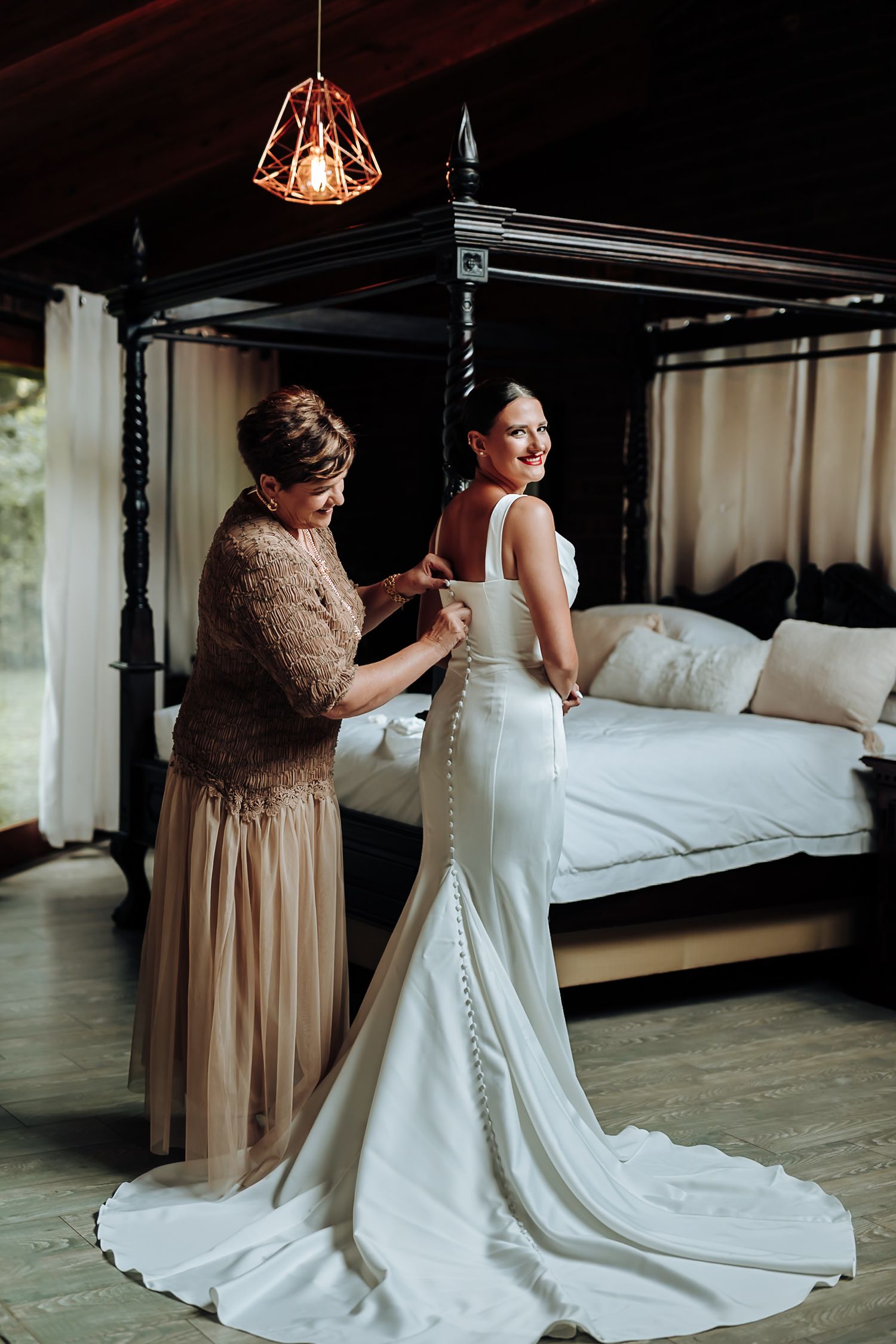 A person in a champagne-colored dress helps another in a white wedding gown with buttons during preparation.