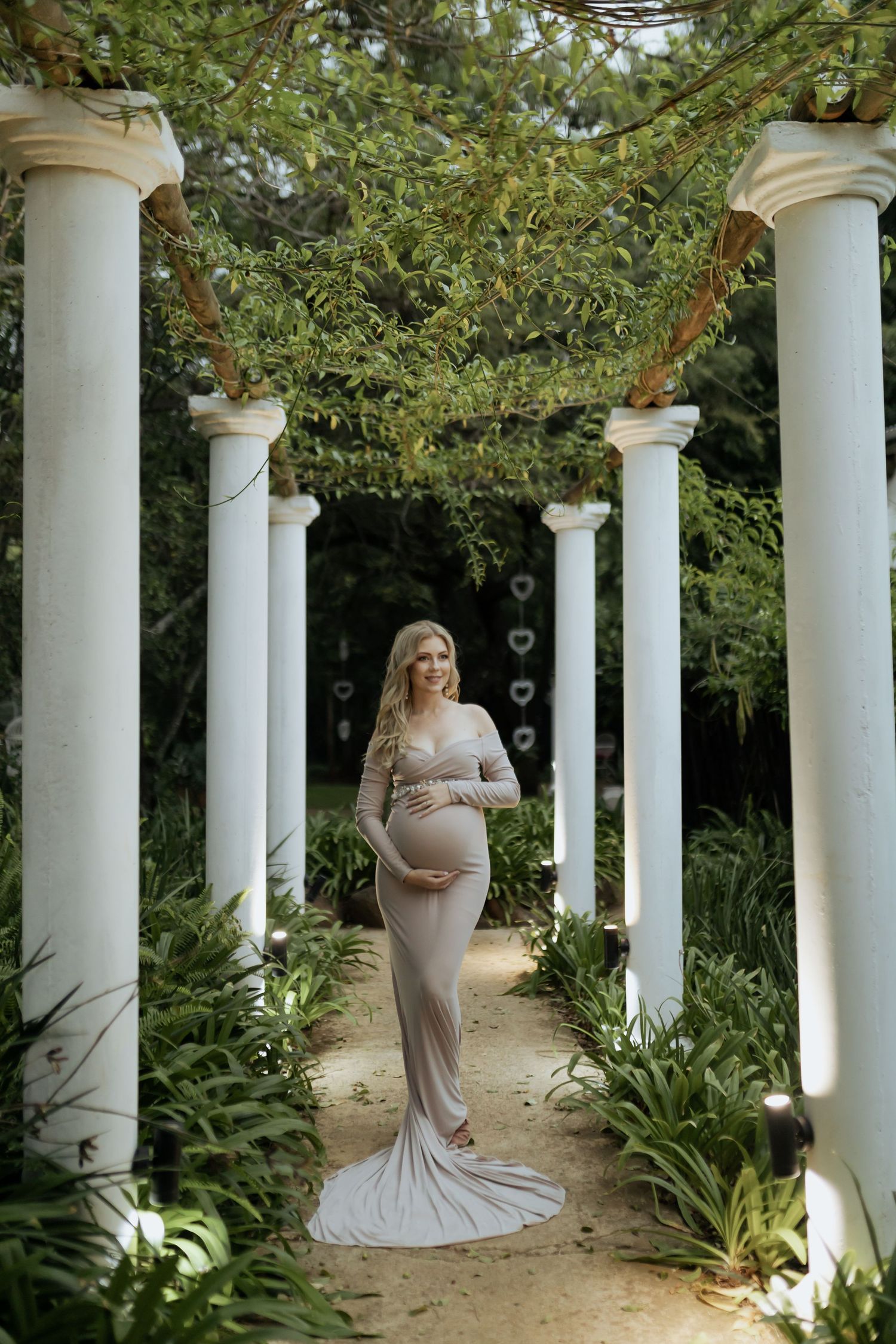 Elegant maternity photoshoot in a garden pergola with white columns and greenery creating a romantic outdoor setting.