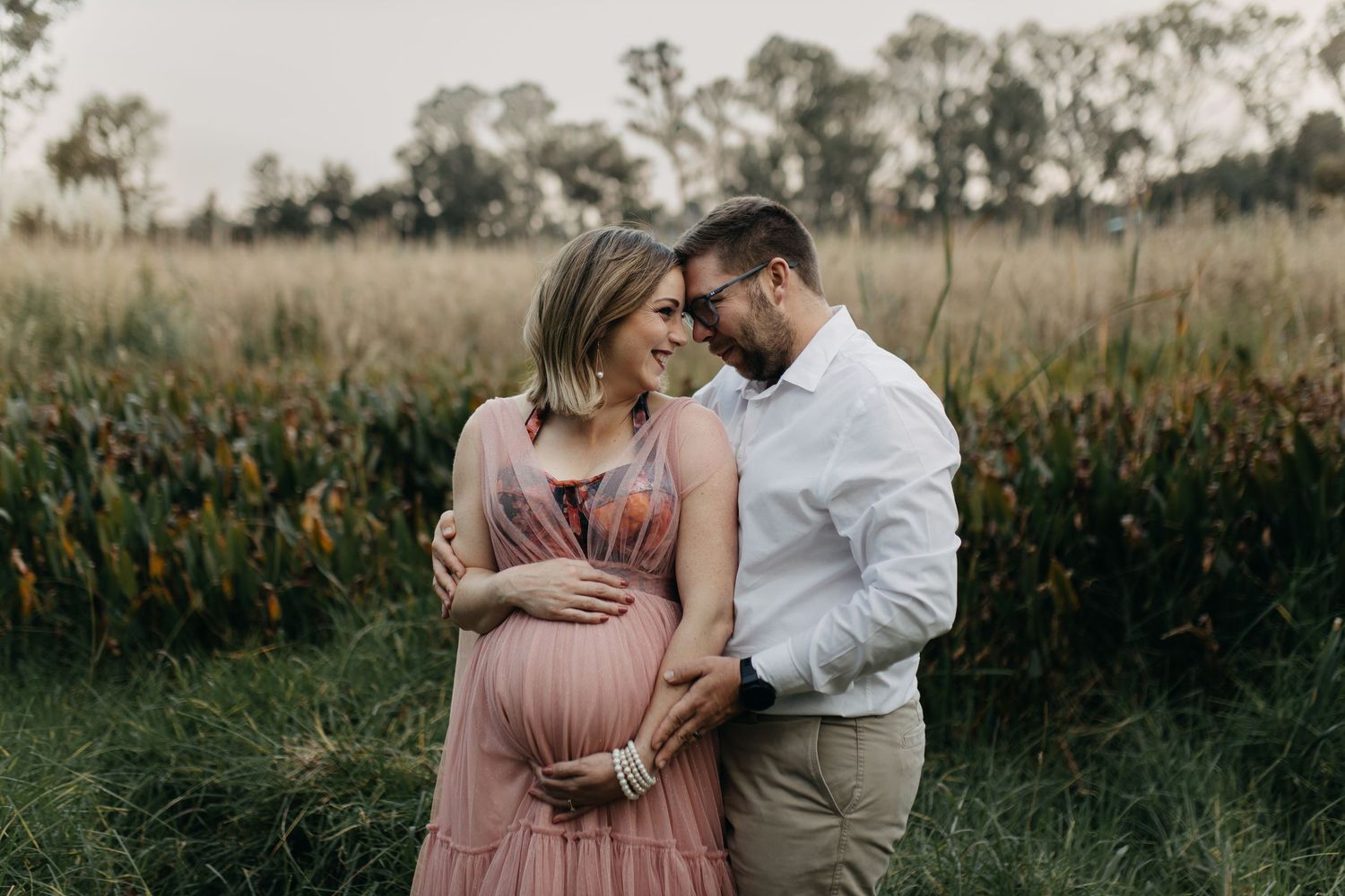 A couple stands together in a rustic field with trees during a romantic golden hour photoshoot.