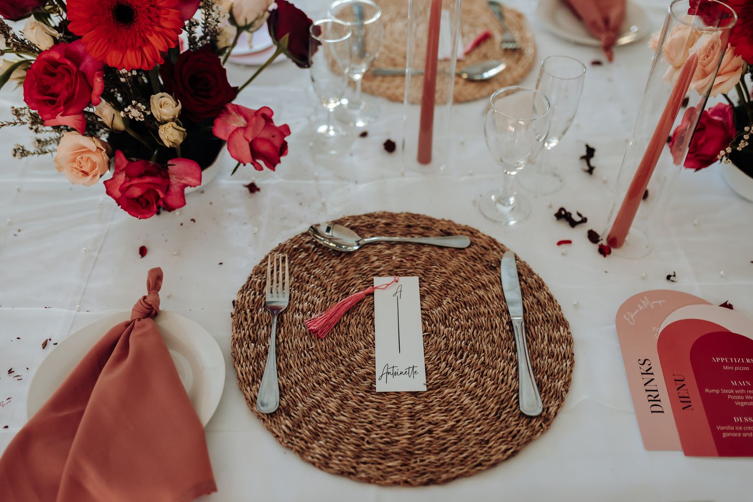 Place setting with woven charger plate, silverware, and pink floral accents on white tablecloth.