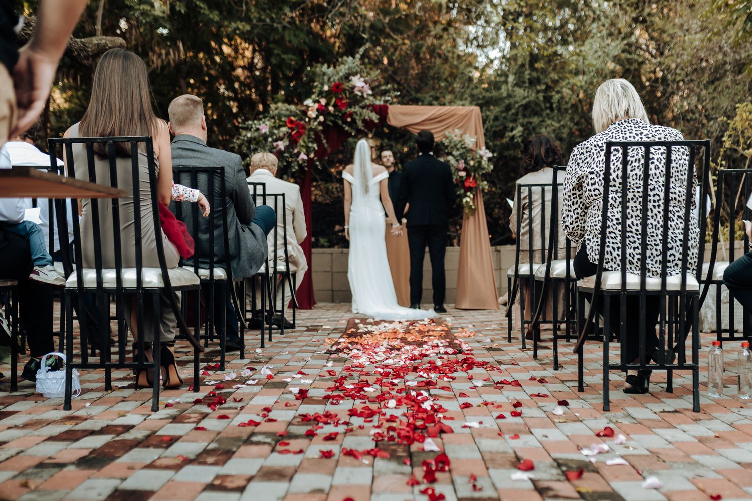 Intimate outdoor wedding ceremony with black chairs and red petals scattered along brick pathway.