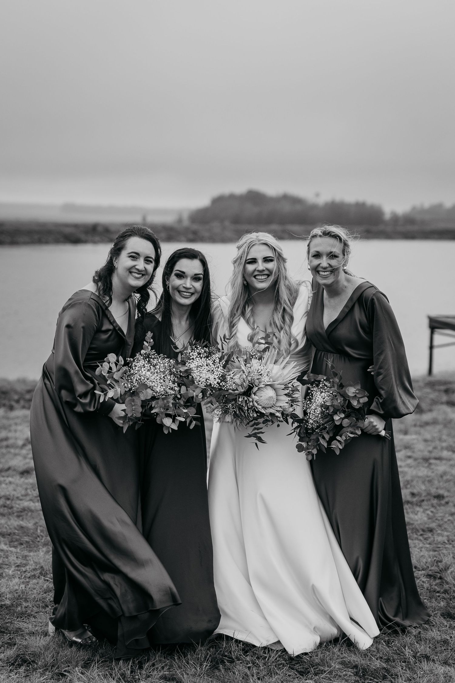 Black and white portrait of bridal party holding bouquets by a lake.
