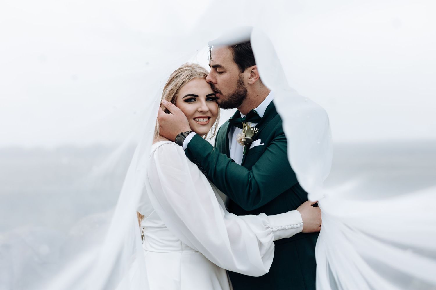 Modern wedding portrait shows couple's intimate embrace with dramatic white veil against cloudy sky.